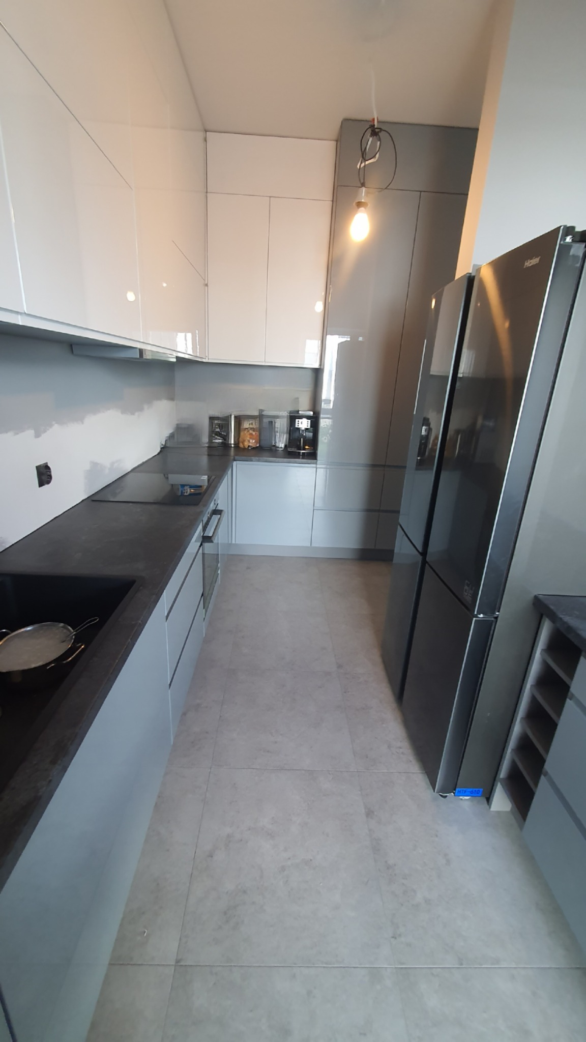 Nowoczesna kuchnia z białymi błyszczącymi szafkami górnymi i szarymi dolnymi, czarny blat, duża lodówka side-by-side, widoczne akcesoria kuchenne na blacie.