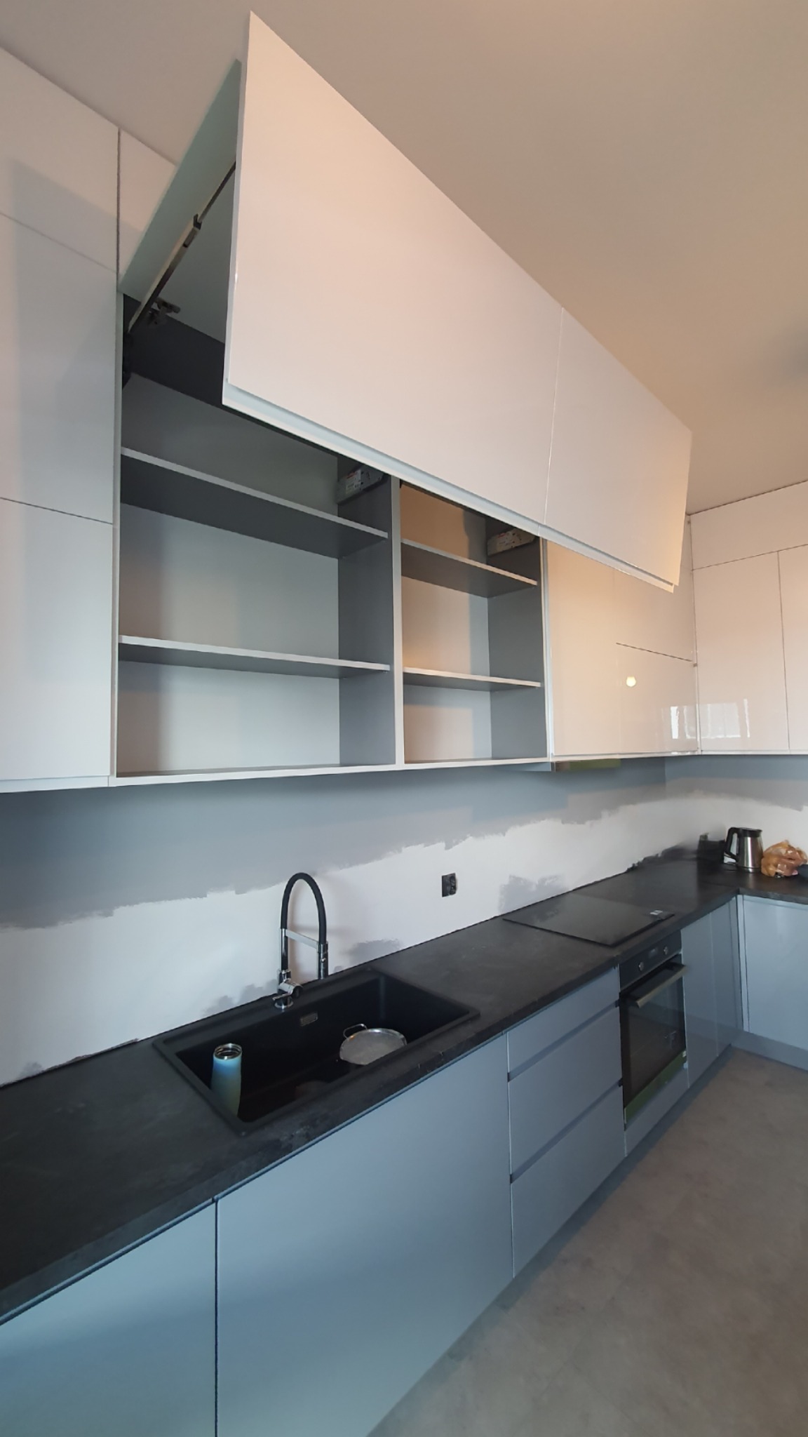 Szara, nowoczesna zabudowa kuchenna z czarnym zlewem i blatem, górne szafki otwarte do góry, widoczne półki, ściana częściowo pomalowana, brak wykończenia.