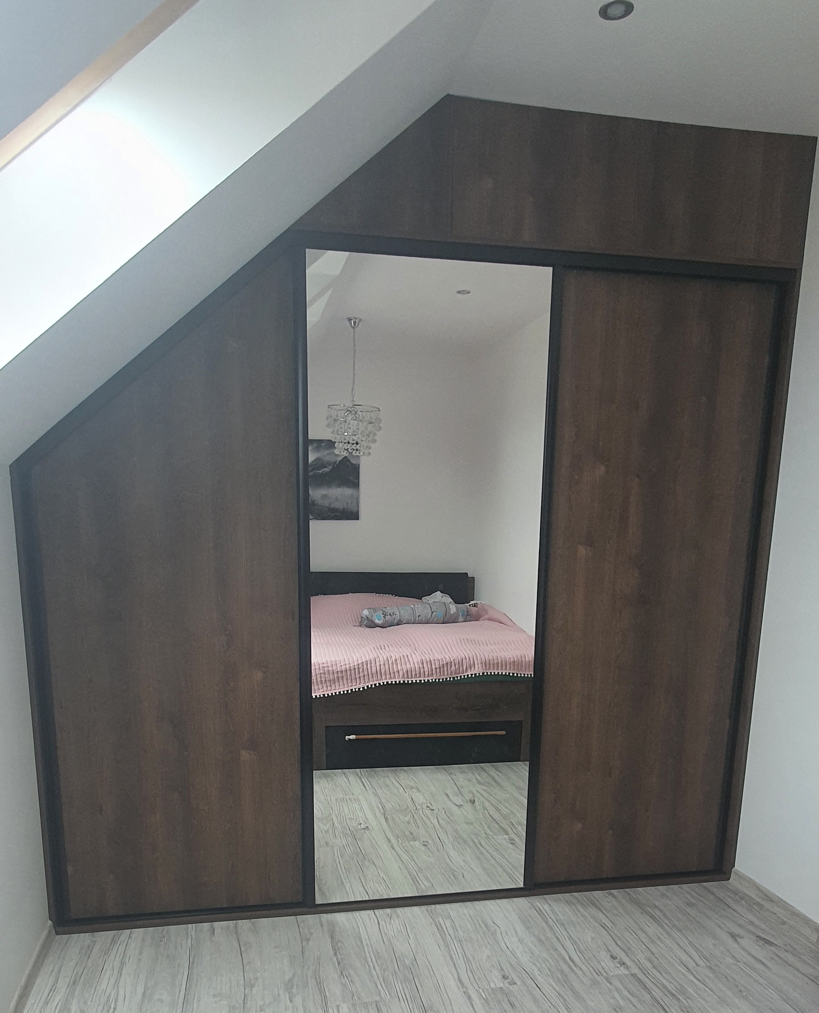 Szafa wnękowa z przesuwnymi drzwiami w kolorze ciemnego drewna, dopasowana do skosu dachu, z lustrem odbijającym sypialnię z łóżkiem i kryształowym żyrandolem.