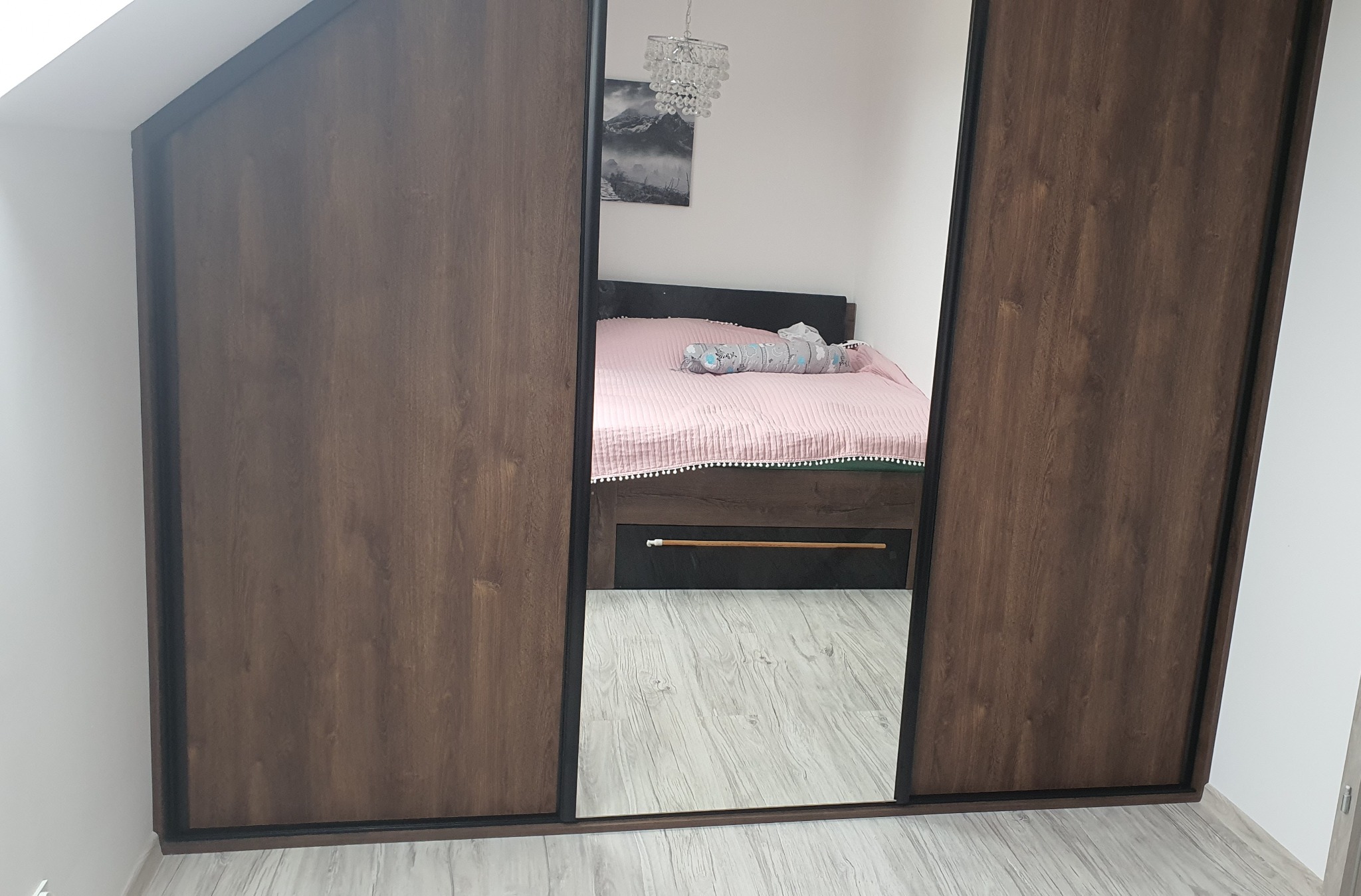 Wnętrze sypialni widoczne w lustrzanych drzwiach przesuwnej szafy w brązowej okleinie drewnopodobnej z czarnymi obramowaniami.