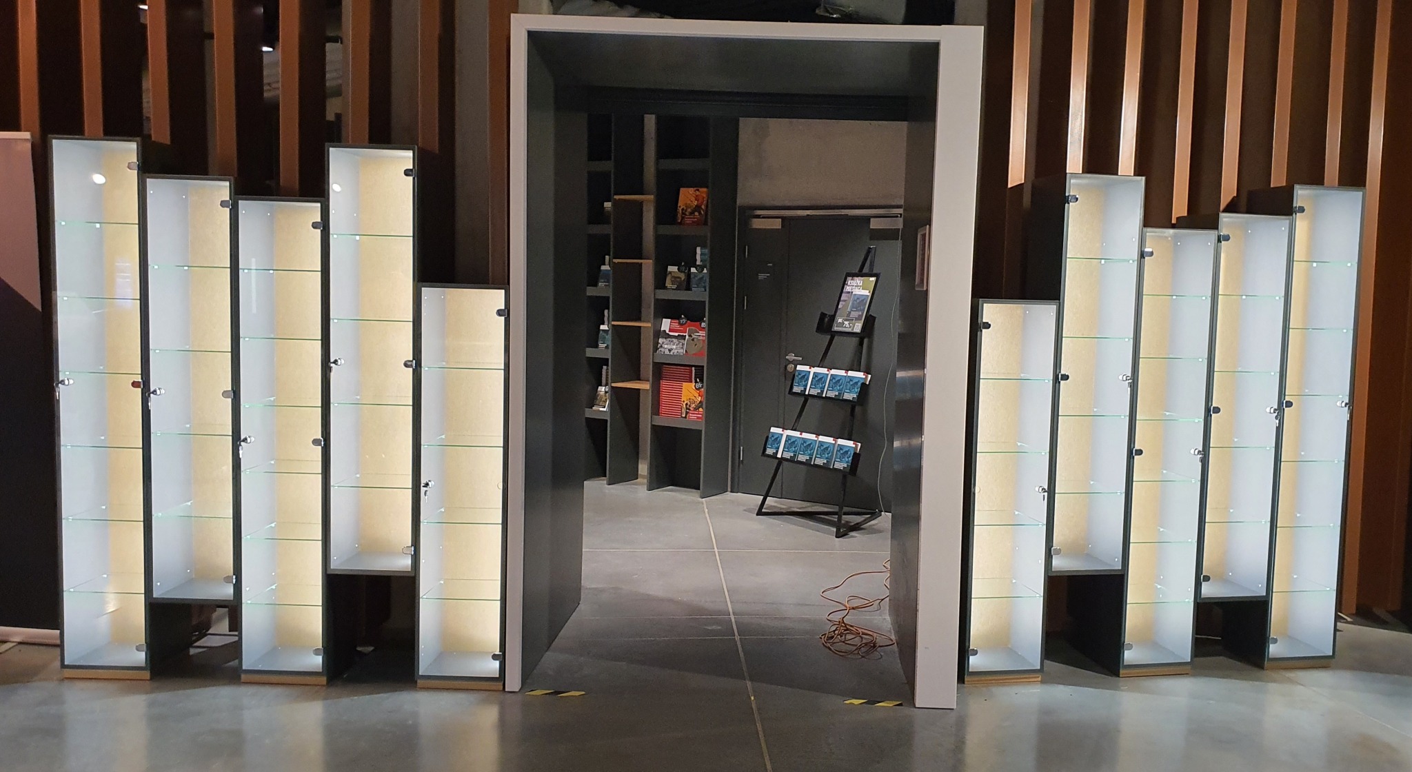 Szereg podświetlanych, przeszklonych witryn ekspozycyjnych o różnej wysokości, ustawionych wzdłuż ściany z drewnianymi panelami, z widokiem na wnętrze sklepu z regałami i stojakiem na ulotki przez...