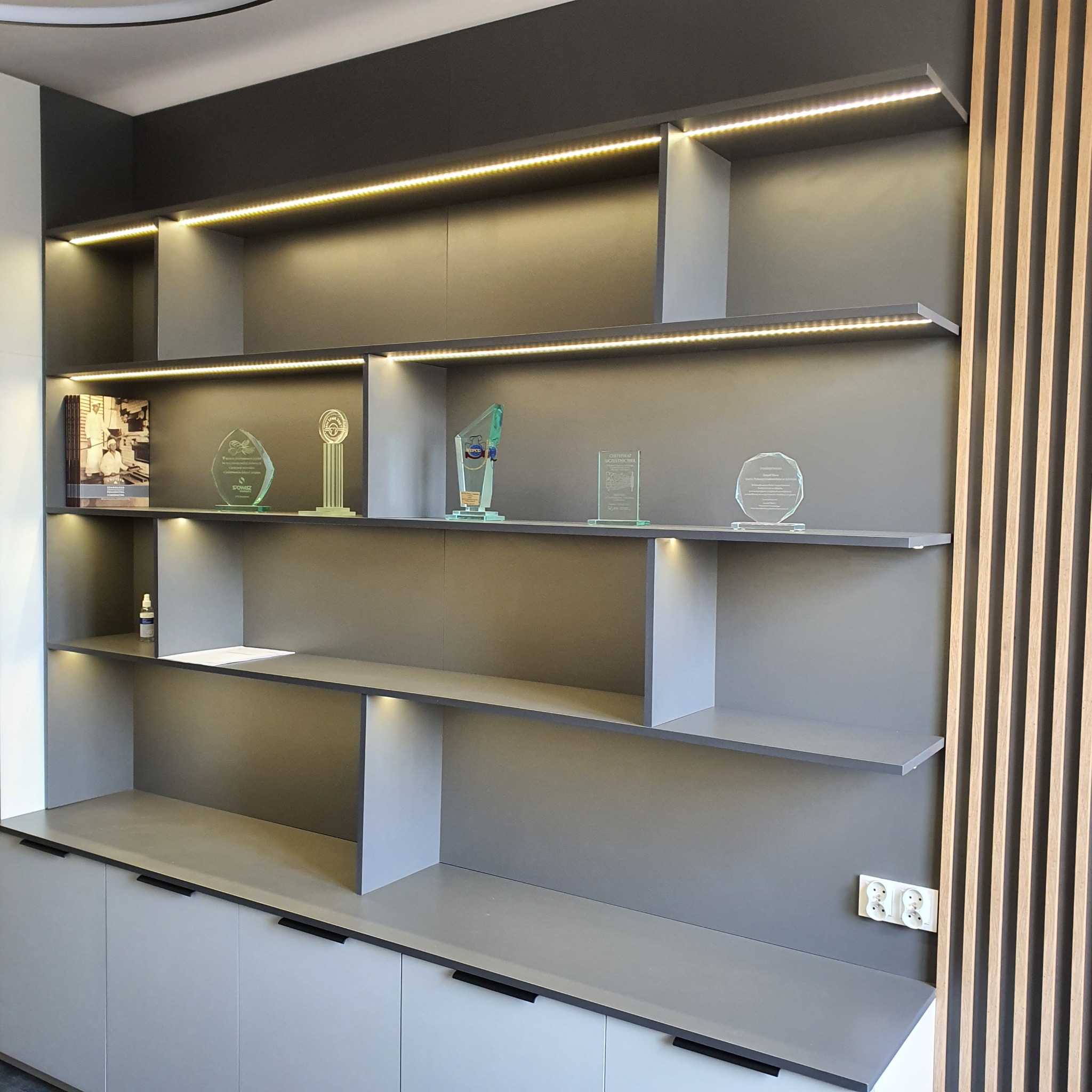 Szara zabudowa meblowa z podświetlonymi półkami, na których stoją książki i nagrody, obok drewniane lamele dekoracyjne i gniazdka elektryczne.