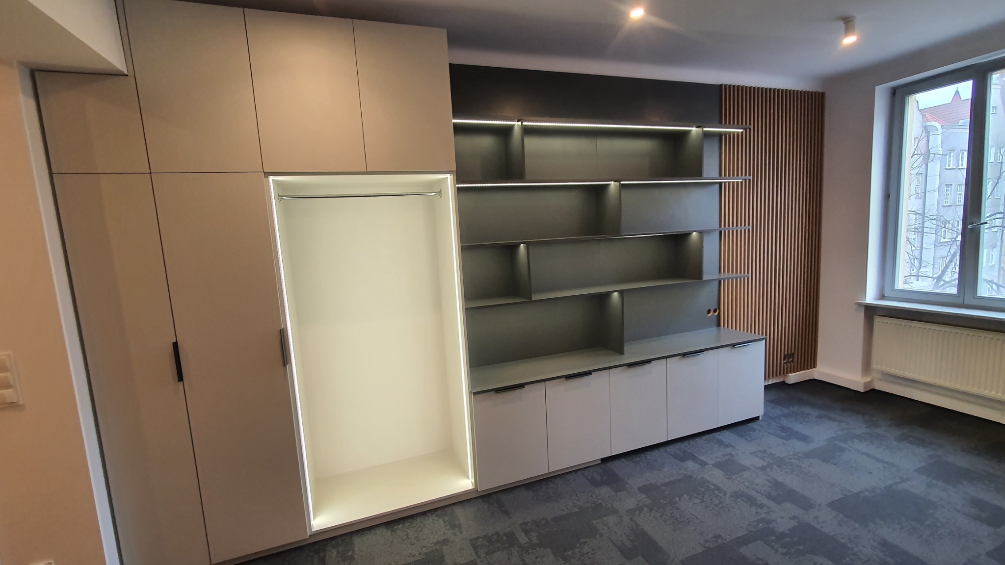 Nowoczesna zabudowa meblowa z szafą z drążkiem na ubrania i regałem z podświetlanymi półkami, uzupełniona o drewniane lamele na ścianie, widoczna na tle okna.