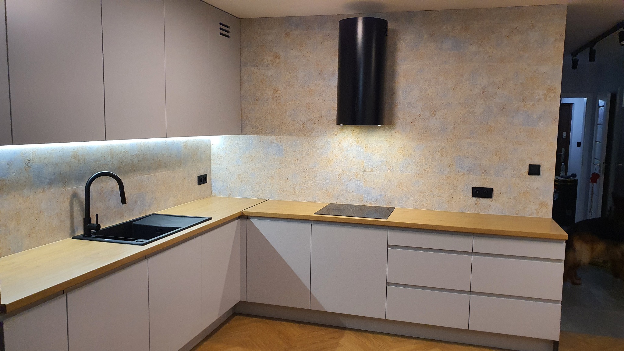 Nowoczesna kuchnia z szarymi szafkami, drewnianym blatem, czarnym zlewem i okrągłym czarnym okapem na tle wzorzystej ściany.