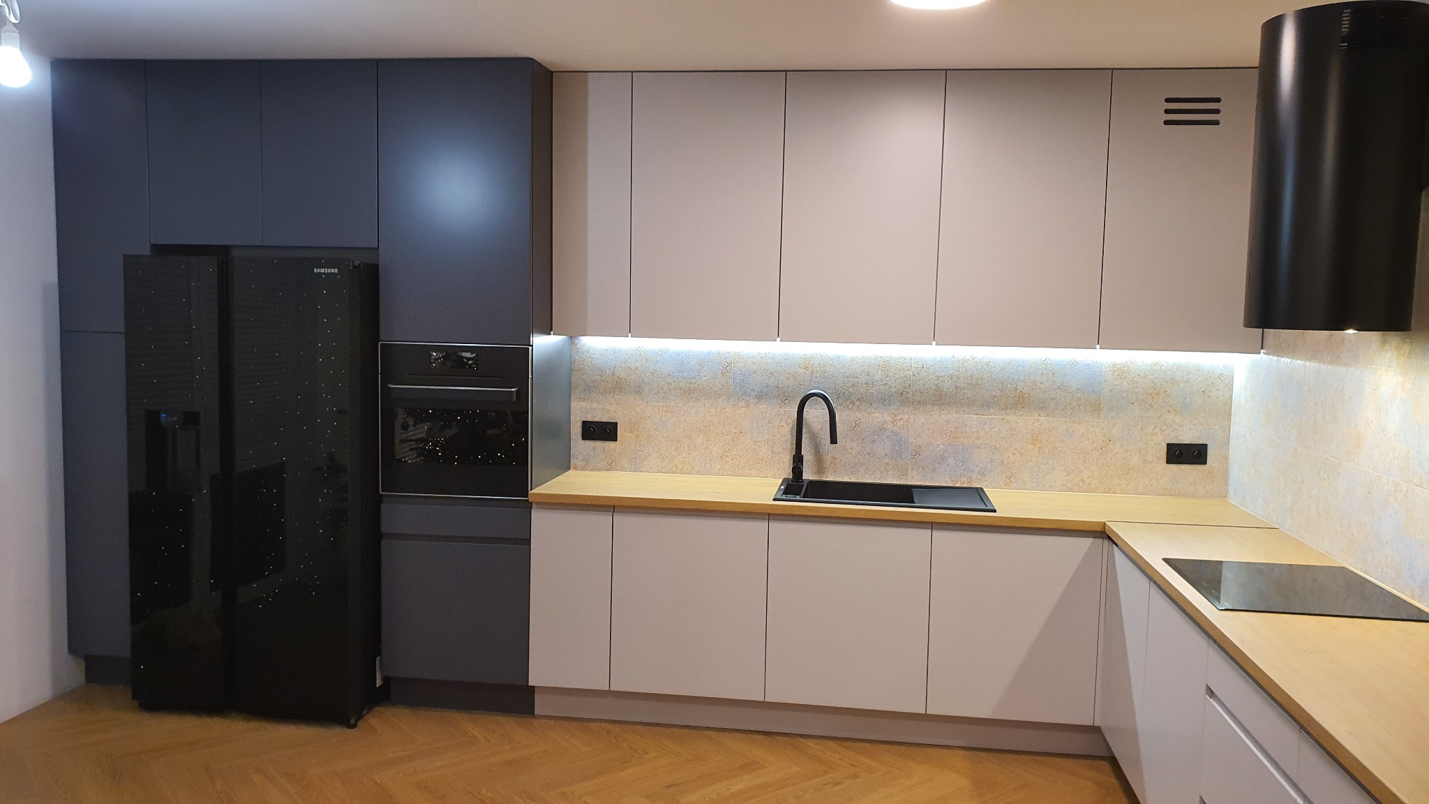 Nowoczesna kuchnia z szarymi i białymi szafkami, czarnym zlewem i sprzętem AGD, oświetleniem LED pod szafkami wiszącymi oraz drewnianym blatem i podłogą z parkietu.