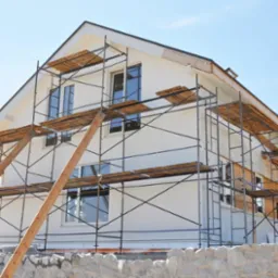 Ocieplany dom jednorodzinny w trakcie prac elewacyjnych, widoczne rusztowania, fragmenty ocieplonej i nieocieplonej ściany, kamienny murek w tle.