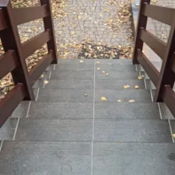 Zewnętrzne schody z szarych płytek z brązową drewnianą balustradą prowadzące do chodnika z kostki brukowej pokrytego opadłymi liśćmi.