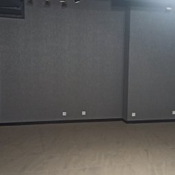 Elizalia Pavlo Tkachuk - Puste wnętrze biura z szarą ścianą o fakturze i widocznymi instalacjami sufitowymi oraz reflektorami punktowymi, podłoga wyłożona płytkami imitującymi marmur.