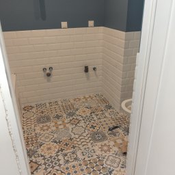 Elizalia Pavlo Tkachuk - Wnętrze łazienki w trakcie remontu: ułożone płytki podłogowe w stylu patchwork, białe płytki ścienne typu metro z ciemnoszarym pasem na górze, wystające rury i instalacje, widoczna część umywalki...
