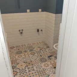 Wnętrze łazienki w trakcie remontu: ułożone płytki podłogowe w stylu patchwork, białe płytki ścienne typu metro z ciemnoszarym pasem na górze, wystające rury i instalacje, widoczna część umywalki...