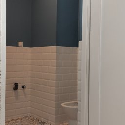 Elizalia Pavlo Tkachuk - Wnętrze łazienki w trakcie remontu: widoczne białe płytki typu metro na ścianach, pomalowana na ciemny kolor górna część ścian, wzorzysta podłoga, biała toaleta i narzędzia budowlane.