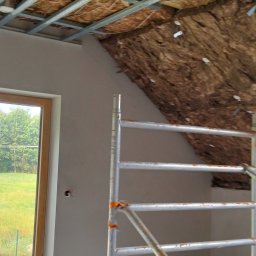 Elizalia Pavlo Tkachuk - Skośne poddasze z oknem, częściowo ocieplone wełną mineralną, widoczne profile sufitowe i rusztowanie. Ściana przy oknie wyszpachlowana.