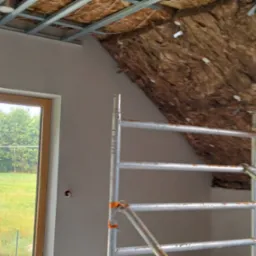 Skośne poddasze z oknem, częściowo ocieplone wełną mineralną, widoczne profile sufitowe i rusztowanie. Ściana przy oknie wyszpachlowana.