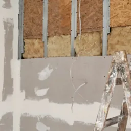 Ściana w trakcie ocieplania: widoczne płyty OSB, wełna mineralna, metalowe profile i częściowo zamontowana płyta karton-gips ze śladami szpachlowania i drabina malarska.