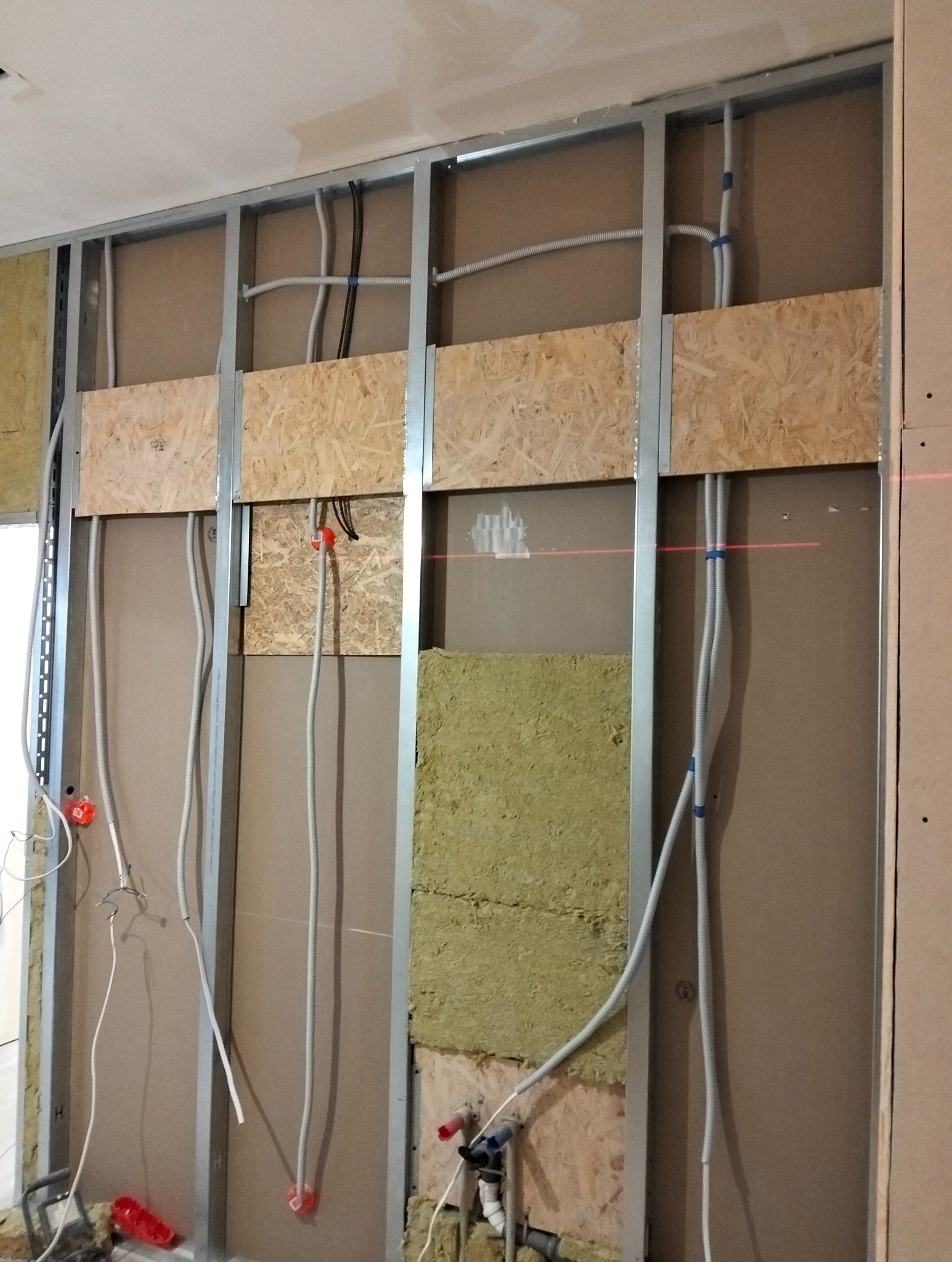 Wnętrze w trakcie remontu: metalowa konstrukcja ścianki działowej z instalacją elektryczną i częściowo zamontowaną izolacją z wełny mineralnej, widoczne przewody i puszki elektryczne, fragment...