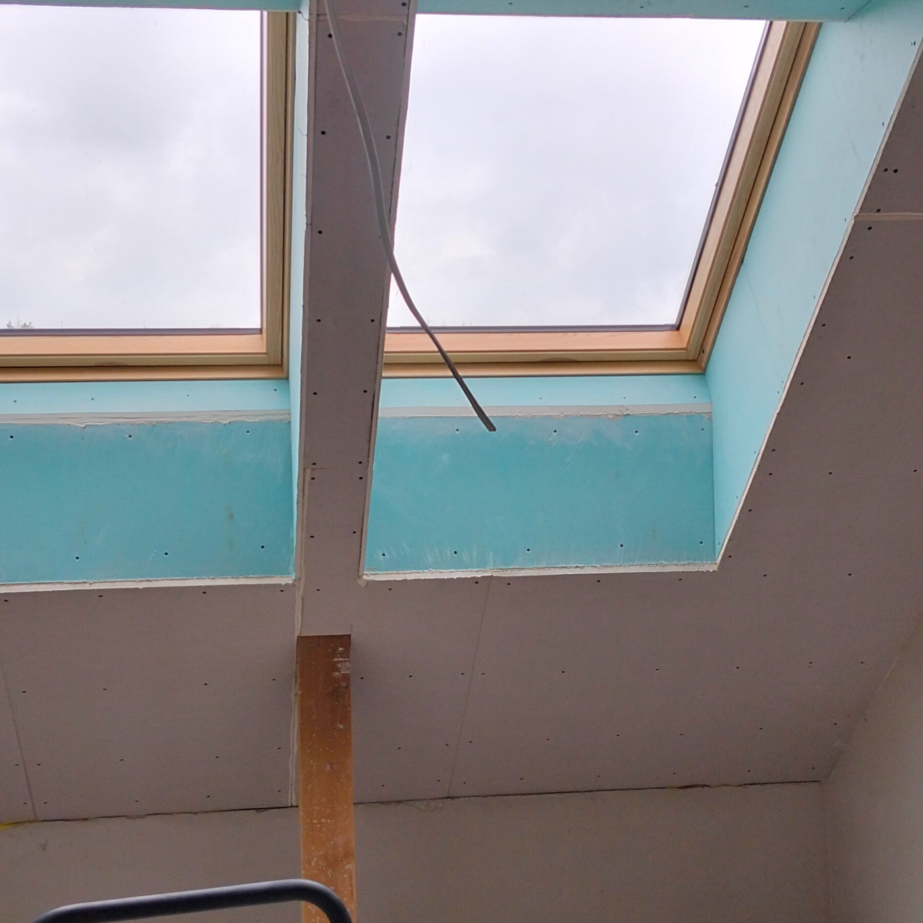 Montaż płyty G-K na suficie poddasza z oknami dachowymi. Widoczne niebieskie płyty i drewniane ramy okien. Kabel zwisa z sufitu. Częściowo widoczna drabina.