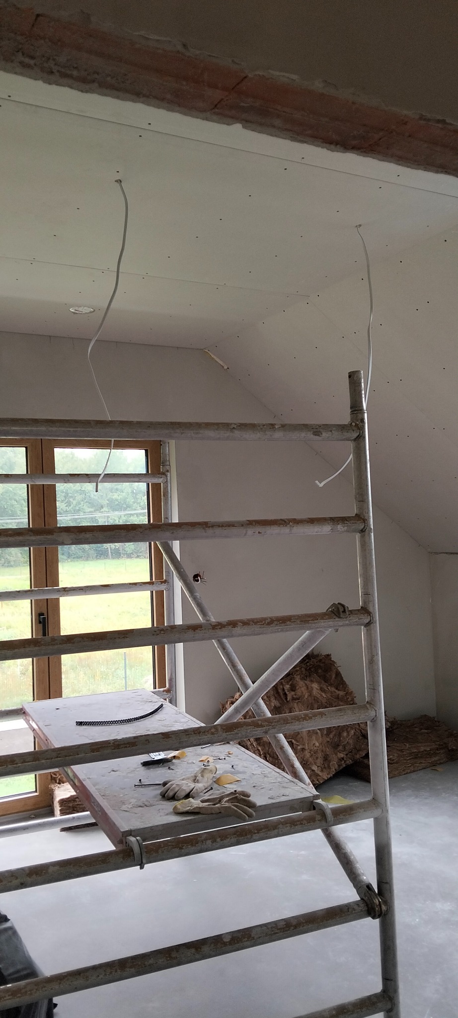 Poddasze po montażu płyt G-K, widoczne kable, rusztowanie z narzędziami i oknem w tle. Izolacja z wełny mineralnej przy ścianie szczytowej.