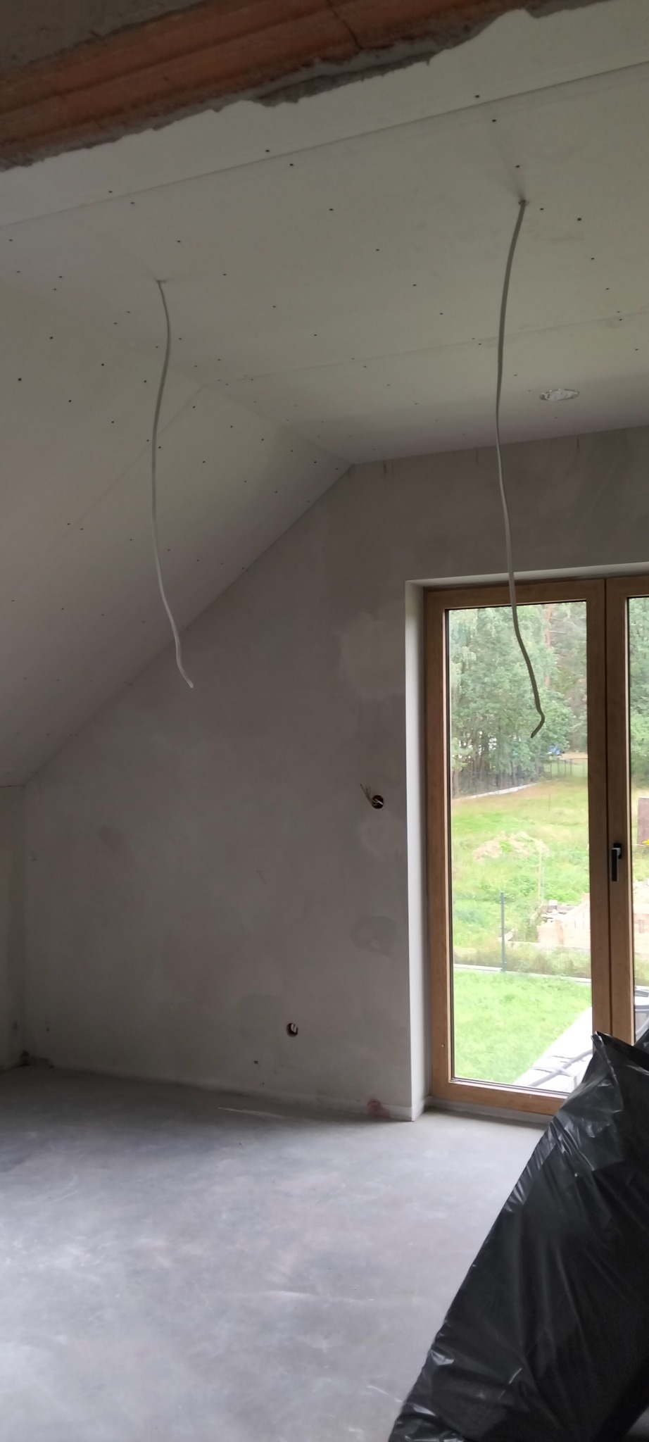 Poddasze po montażu płyt gipsowo-kartonowych, z widocznymi przewodami elektrycznymi i oknem z widokiem na zieleń. Ściany przygotowane do dalszych prac wykończeniowych.