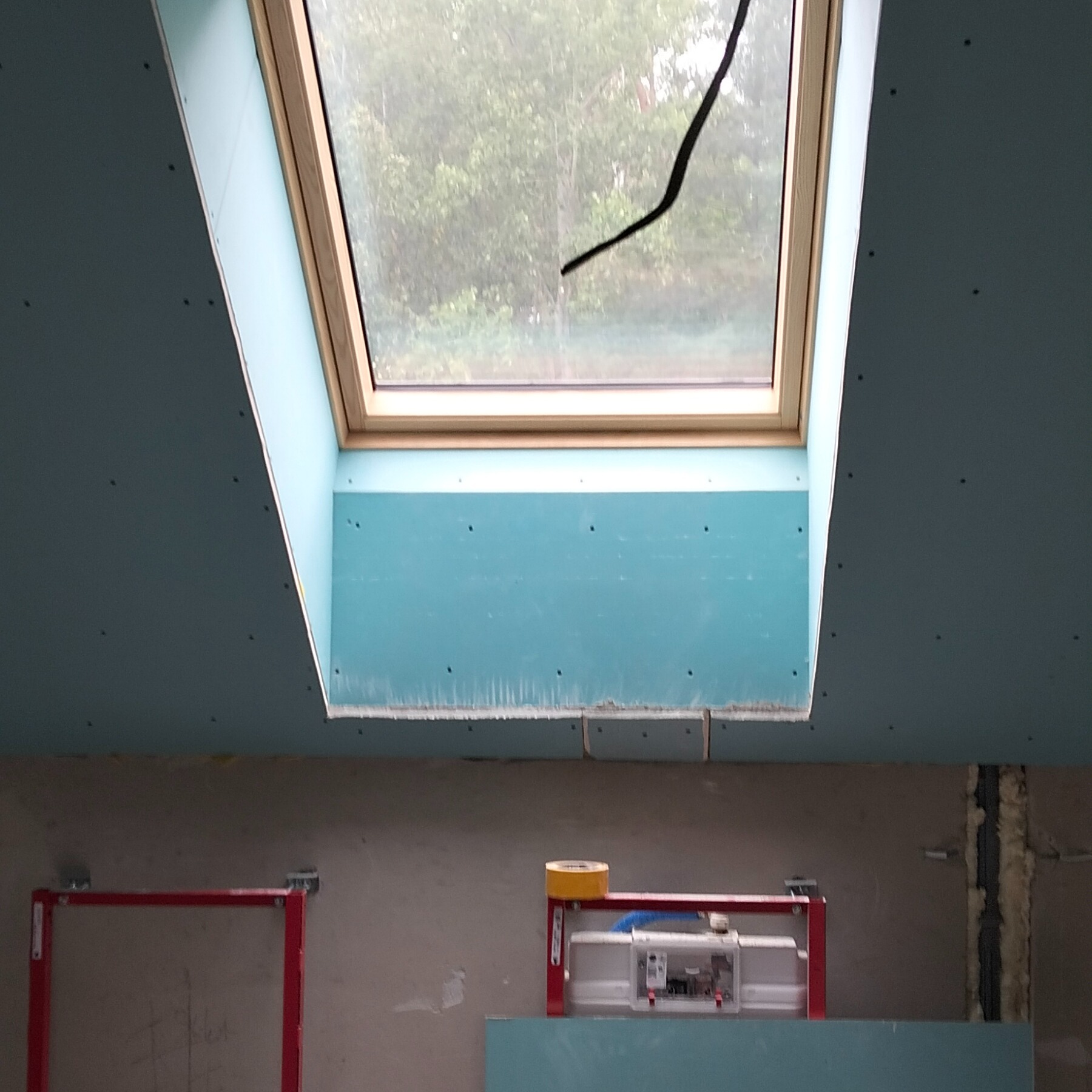 Montaż okna dachowego z płyt kartonowo-gipsowych, widoczne łączenia i fragment instalacji elektrycznej. Widok na zewnątrz przez okno.