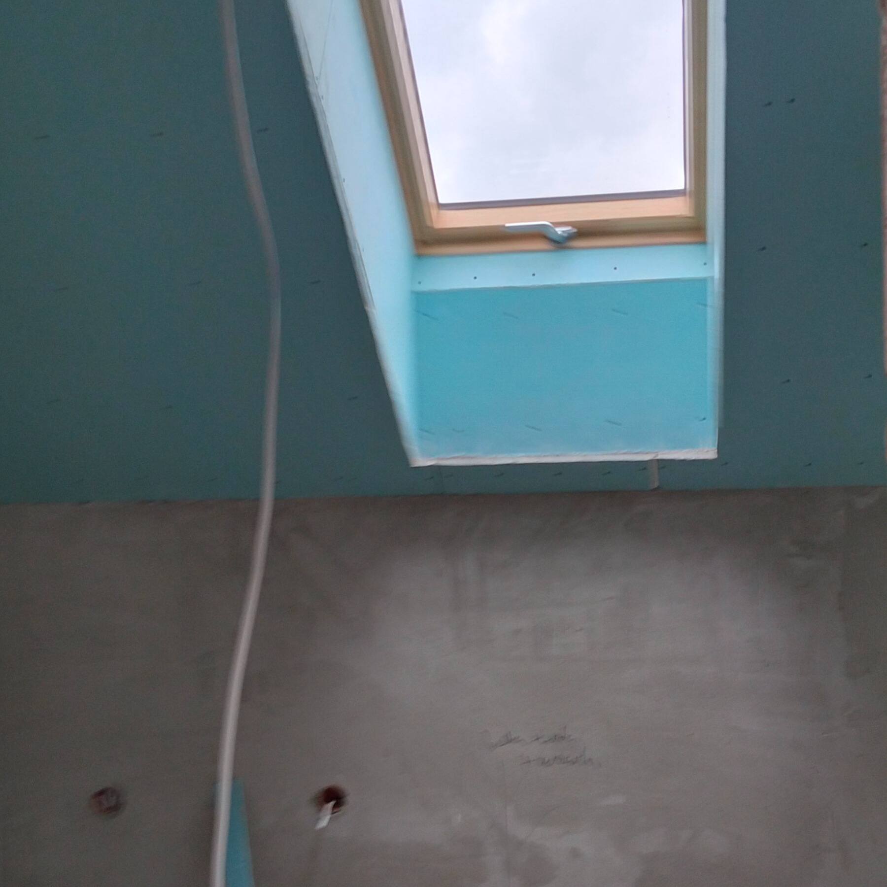 Montaż okna dachowego w poddaszu z płytami GK w kolorze błękitnym. Widoczne przewody elektryczne i surowa ściana. Ujęcie od dołu, z perspektywą na okno.