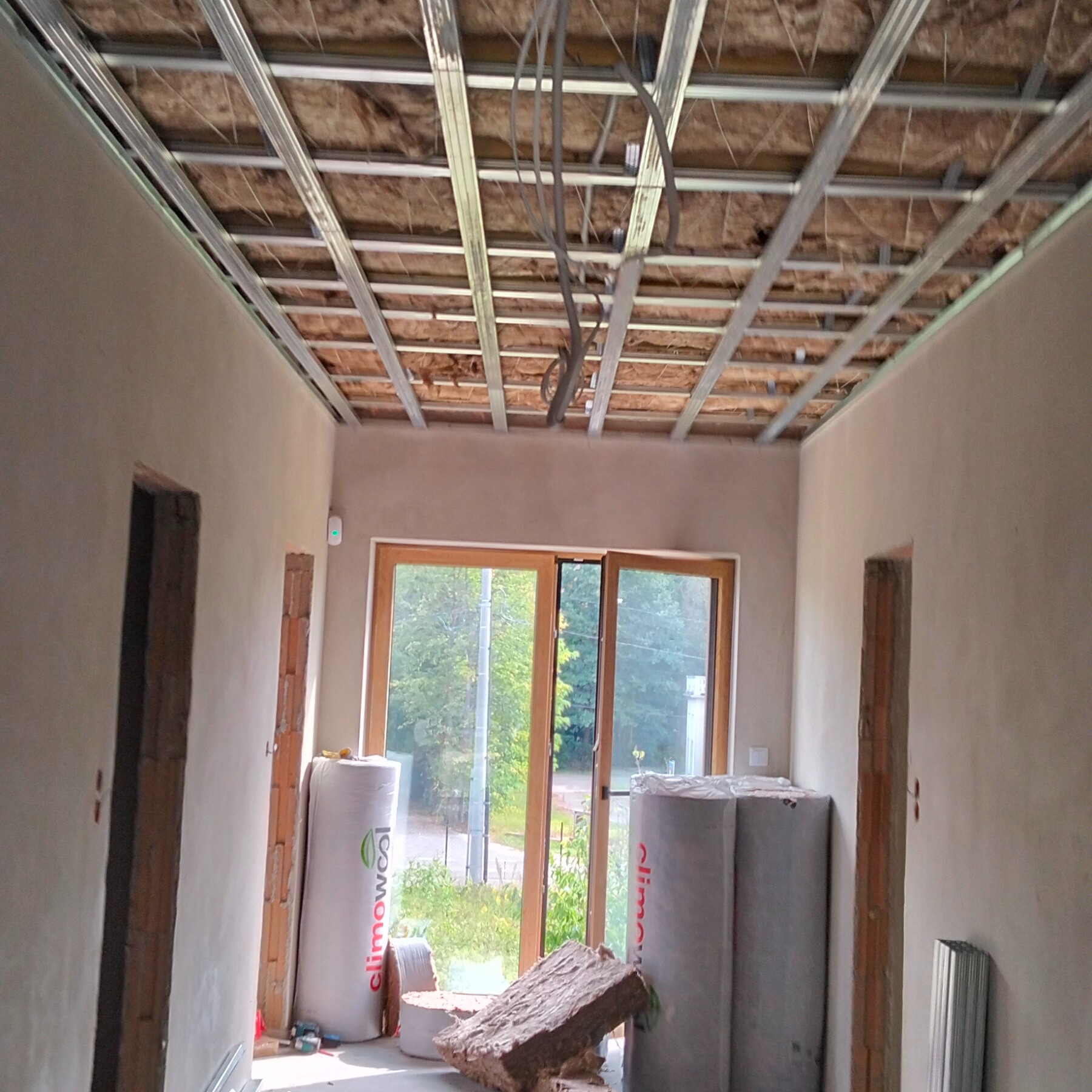 Montaż sufitu podwieszanego z izolacją w Pruszkowie. Widoczne profile, wełna mineralna i częściowo wykończone ściany. Okno w tle. Prace wykończeniowe w toku.