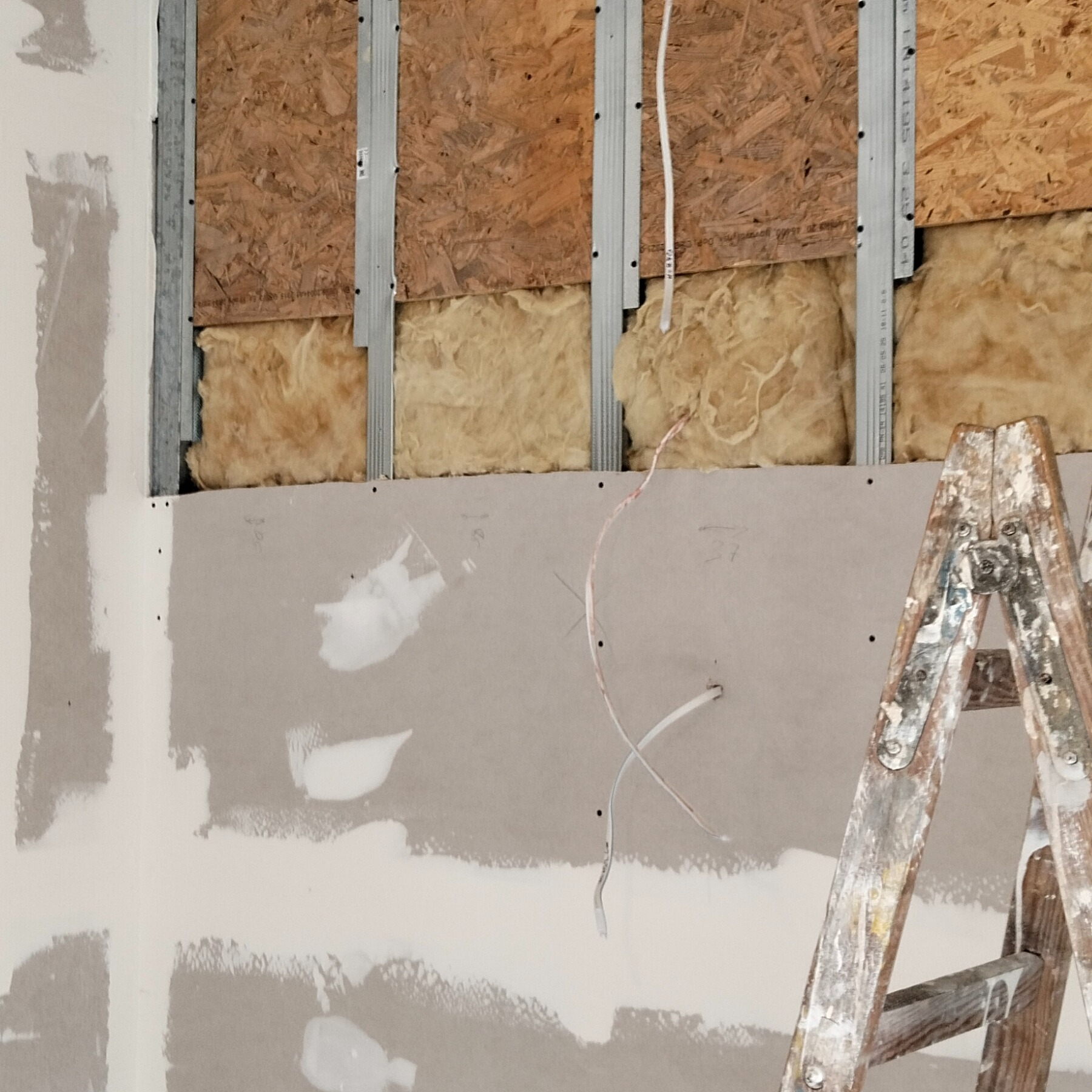 Ściana w trakcie ocieplania: widoczne płyty OSB, wełna mineralna, metalowe profile i częściowo zamontowana płyta karton-gips ze śladami szpachlowania i drabina malarska.