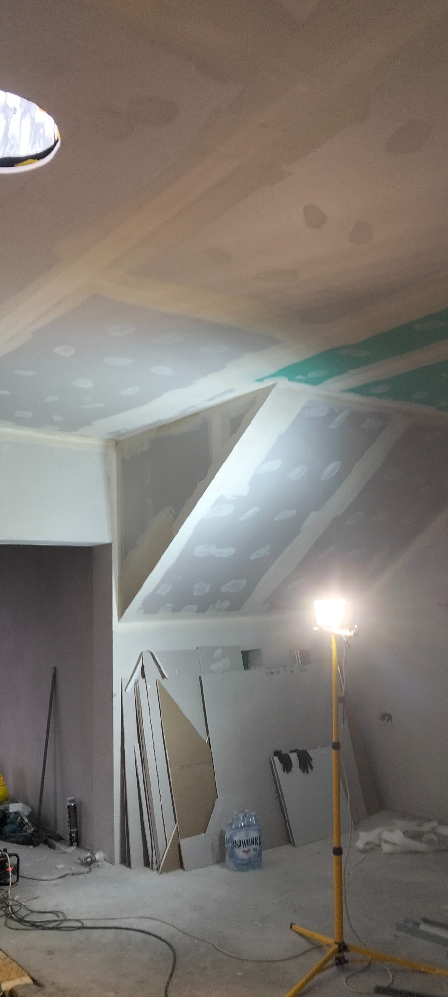 Poddasze w trakcie wykańczania: widoczne płyty karton-gips na skosach, szpachlowane łączenia, lampa budowlana i materiały budowlane na podłodze.