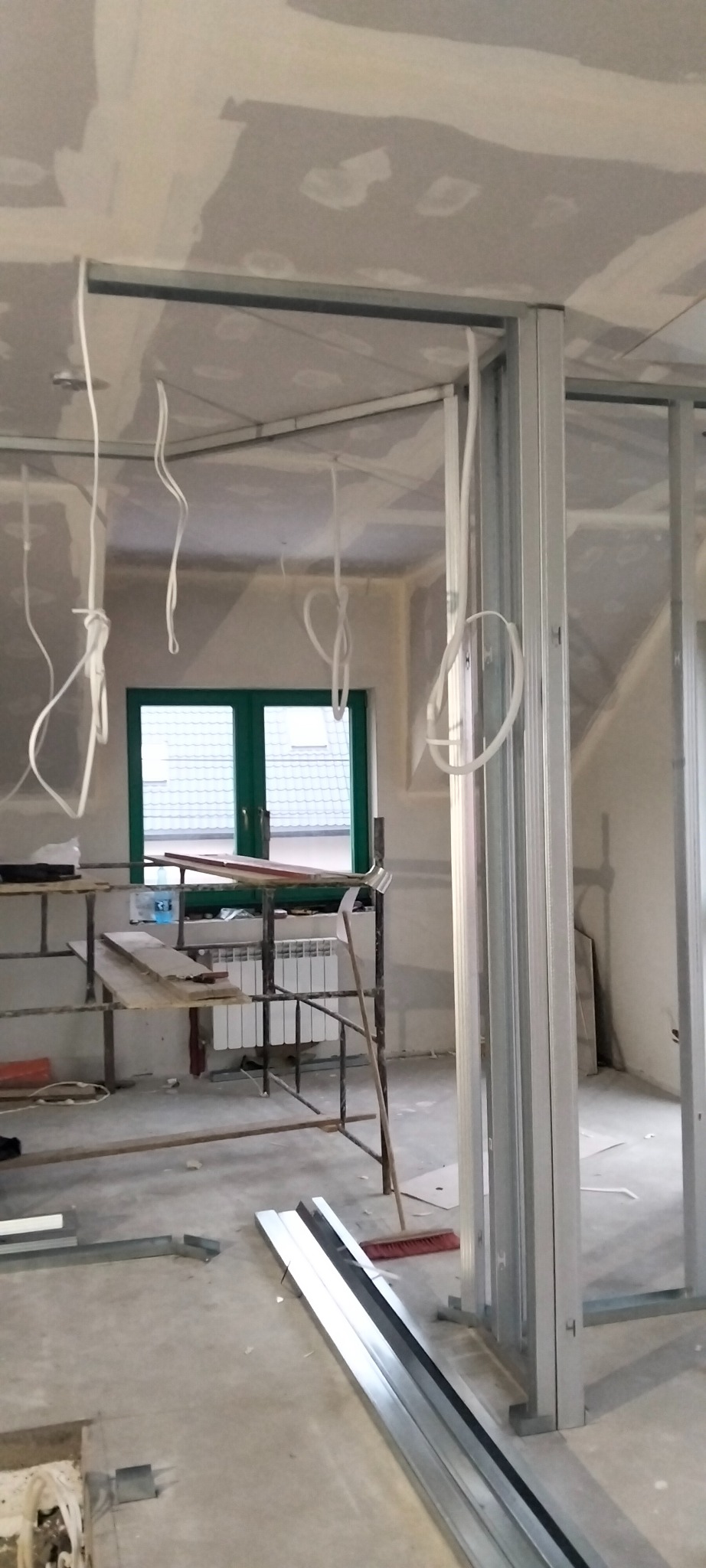 Wnętrze w trakcie prac: metalowa konstrukcja ścian działowych, częściowo wykończona płyta gipsowa, kable zwisające z sufitu, rusztowanie i okno w tle.