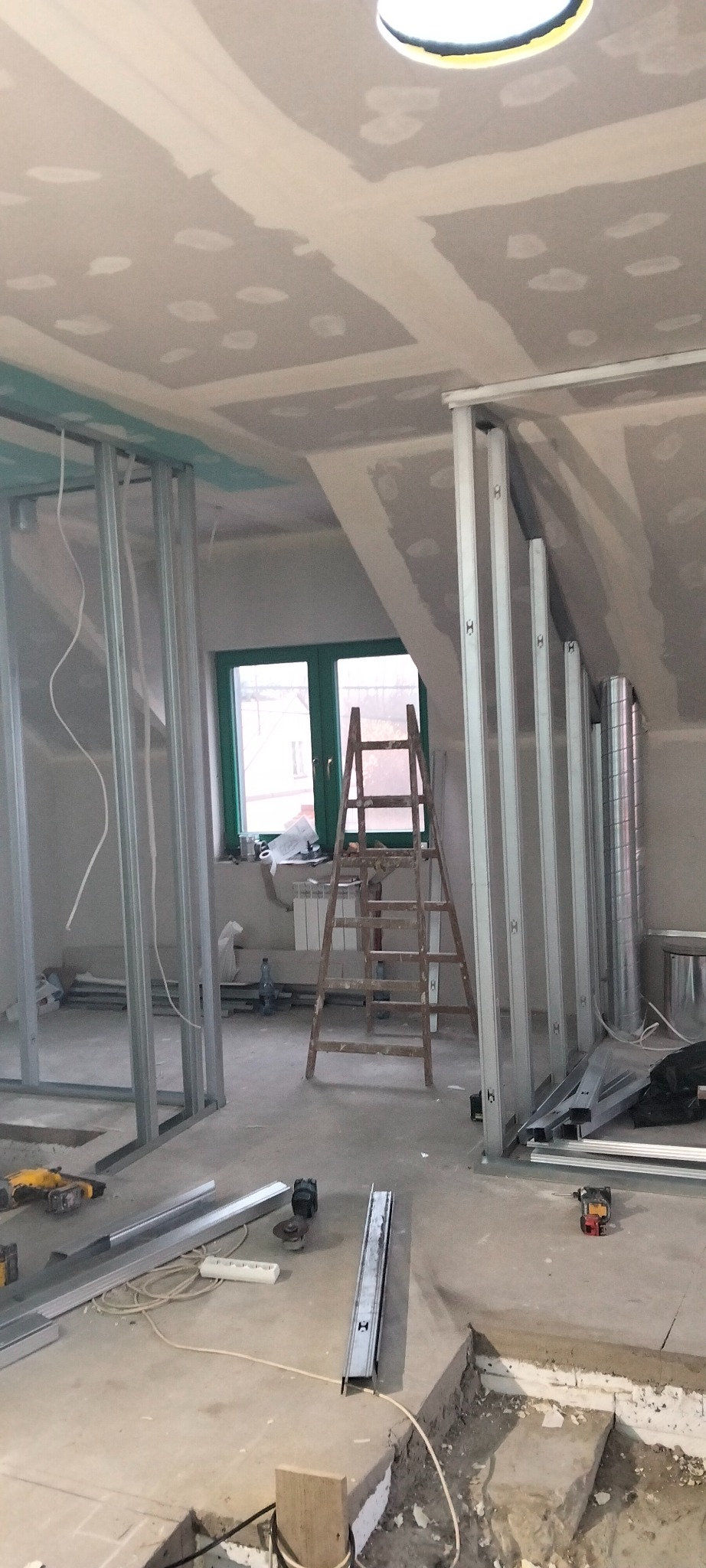 Poddasze w trakcie adaptacji: metalowe profile ścianek działowych, częściowo zamontowane płyty gipsowe na skosach, drabina i narzędzia na podłodze. Widok na okno.