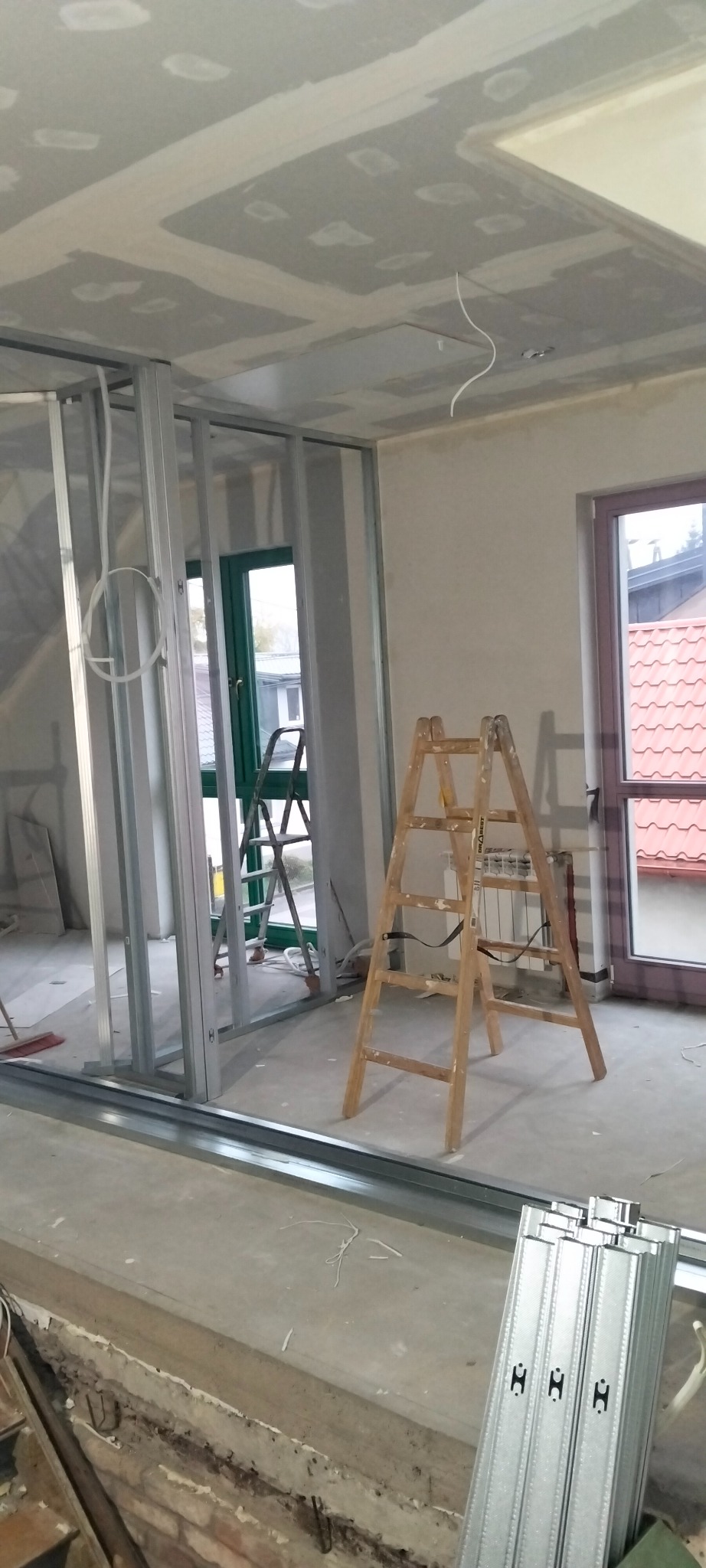 Wnętrze w trakcie prac: metalowa konstrukcja ścian działowych, częściowo zamontowane płyty gipsowe na suficie, drabina drewniana, okno z widokiem na zewnątrz.