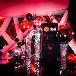 MIRANDA EVENTS - Event z robotem podającym prezenty DPD, dekoracje z balonów i błyszczących elementów. Mężczyzna w okularach obserwuje. Czerwone oświetlenie tworzy klubową atmosferę.
