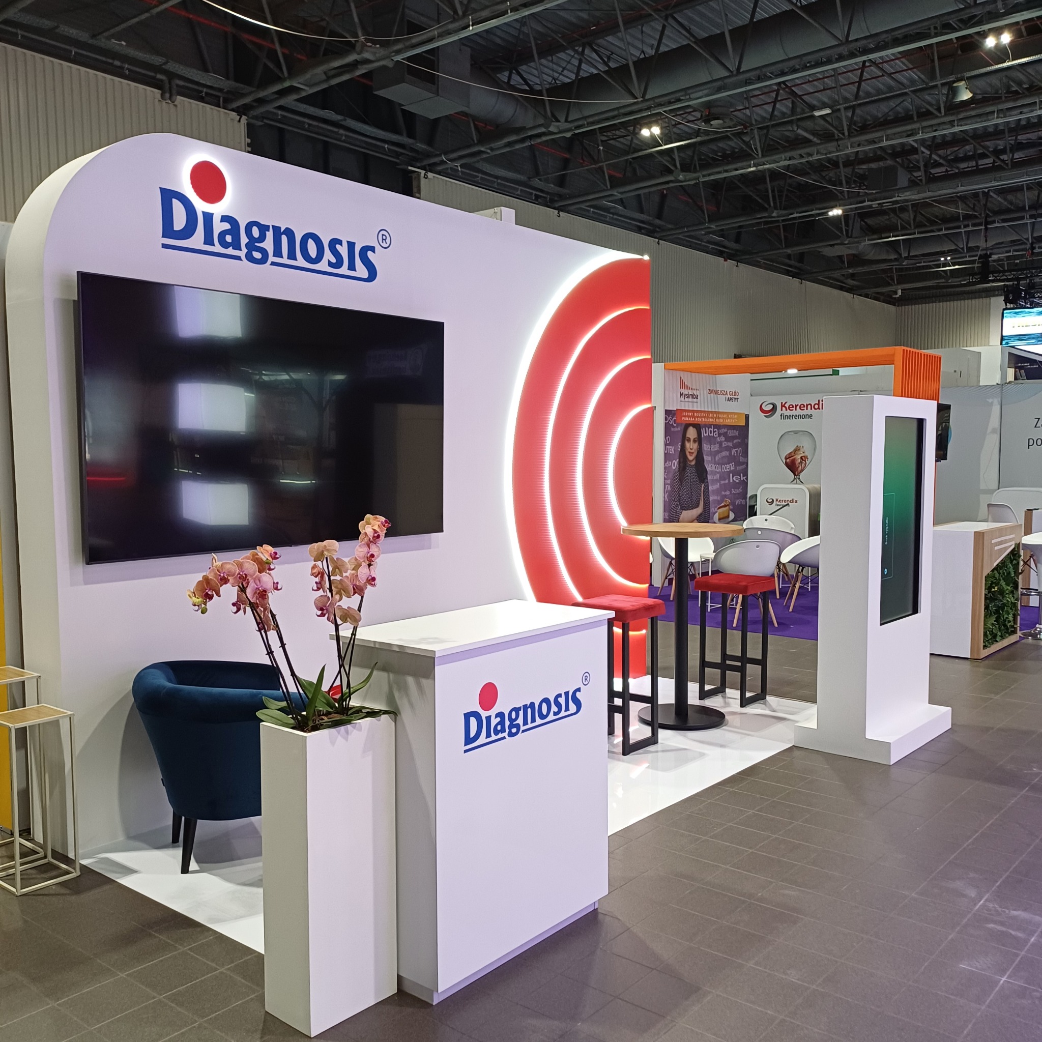 Stoisko targowe firmy Diagnosis S.A. z dotykowym totemem multimedialnym, ekranem, stolikiem barowym z krzesłami, oraz dekoracją z kwiatów orchidei w białej donicy. W tle widoczne inne stoiska.