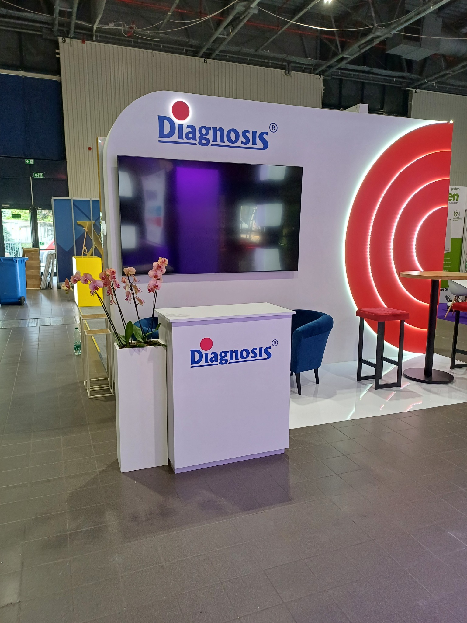 Stoisko targowe z logo firmy Diagnosis, wyposażone w ekran, ladę recepcyjną, krzesło i stolik barowy, ozdobione kwiatami w białej donicy.
