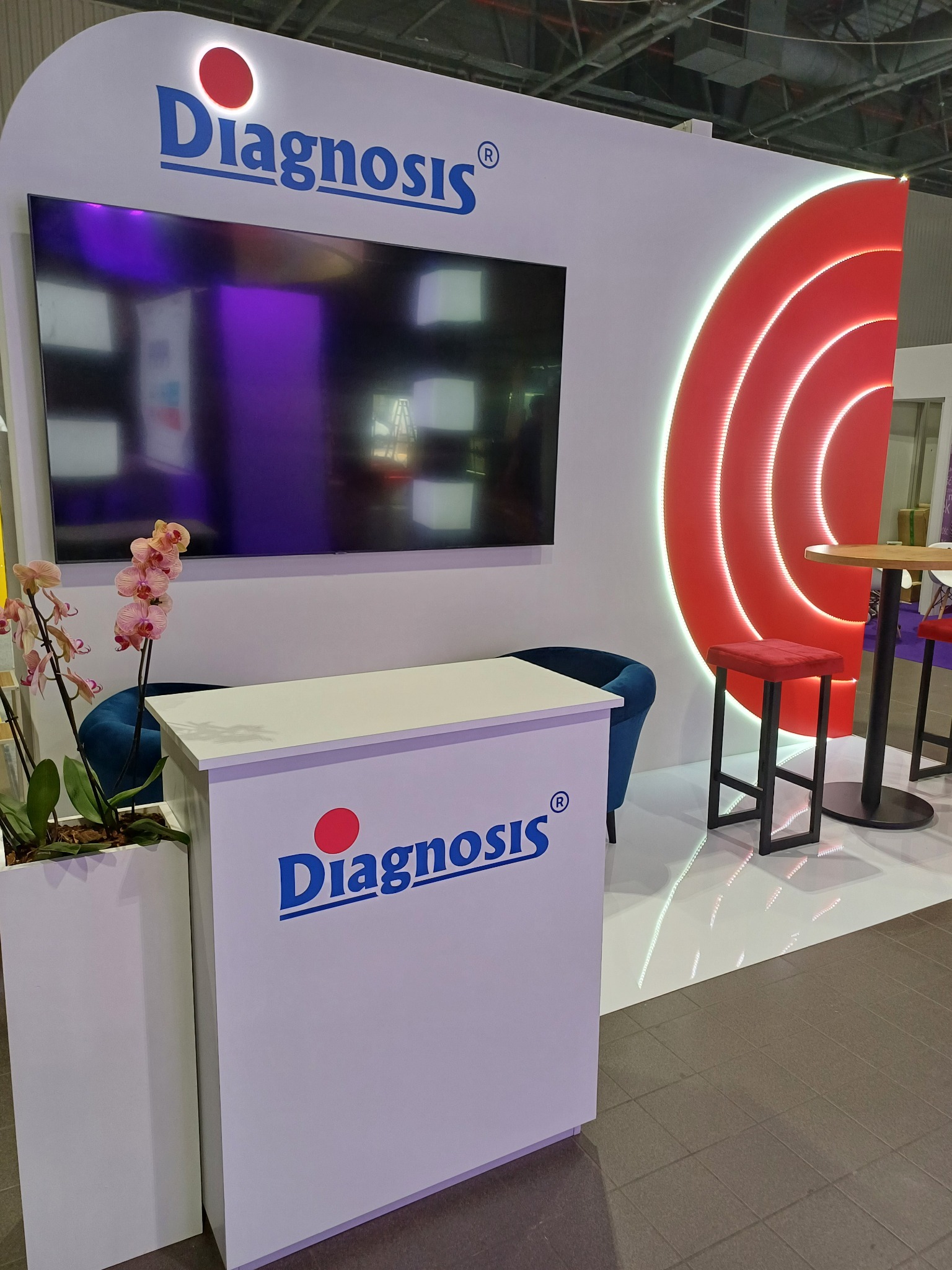 Nowoczesne stoisko targowe z podświetlanym logotypem, dużym ekranem, kwiatami orchidei i czerwonymi akcentami w postaci krzeseł i dekoracji ściennej.
