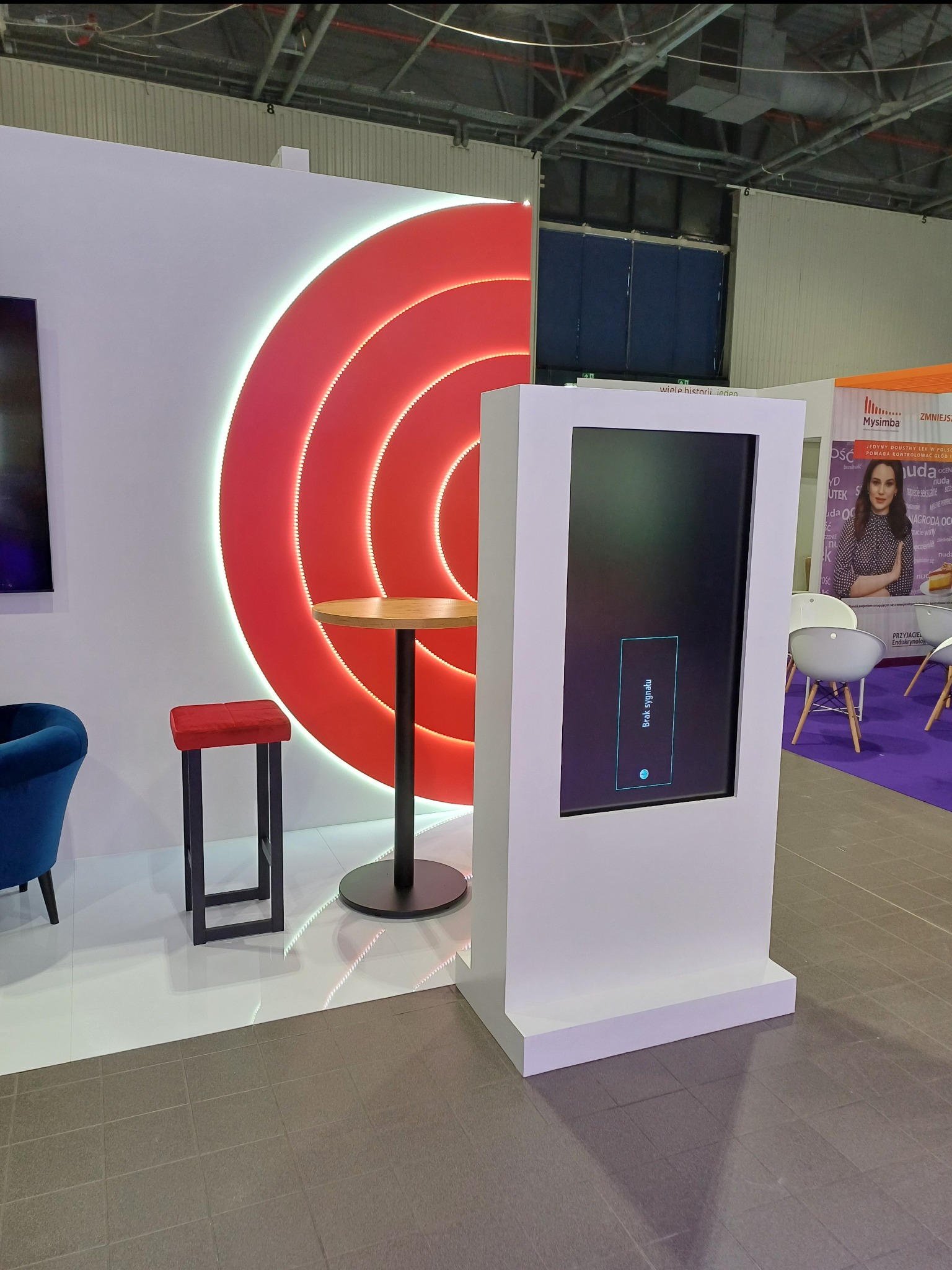 Aranżacja stoiska targowego z podświetlanym czerwonym panelem w kształcie okręgów, białym panelem z ekranem wyświetlającym komunikat 'Brak sygnału', stolikiem barowym i krzesłem w kolorze czerwonym...