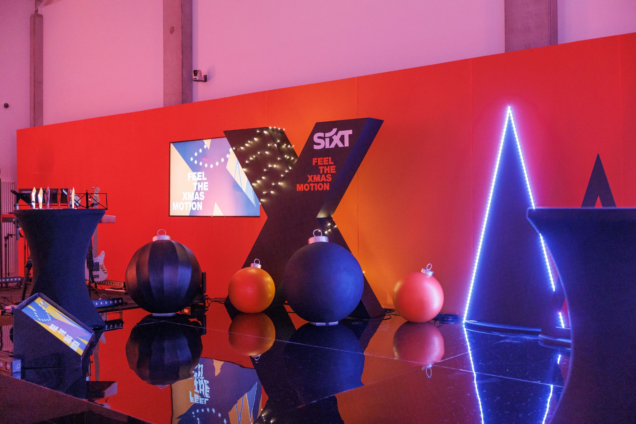 Scena eventowa z dekoracjami świątecznymi: podświetlony neonowy trójkąt, logo firmy SIxT w kształcie litery X z lampkami, oraz duże bombki w kolorach czarnym i pomarańczowym, ustawione...