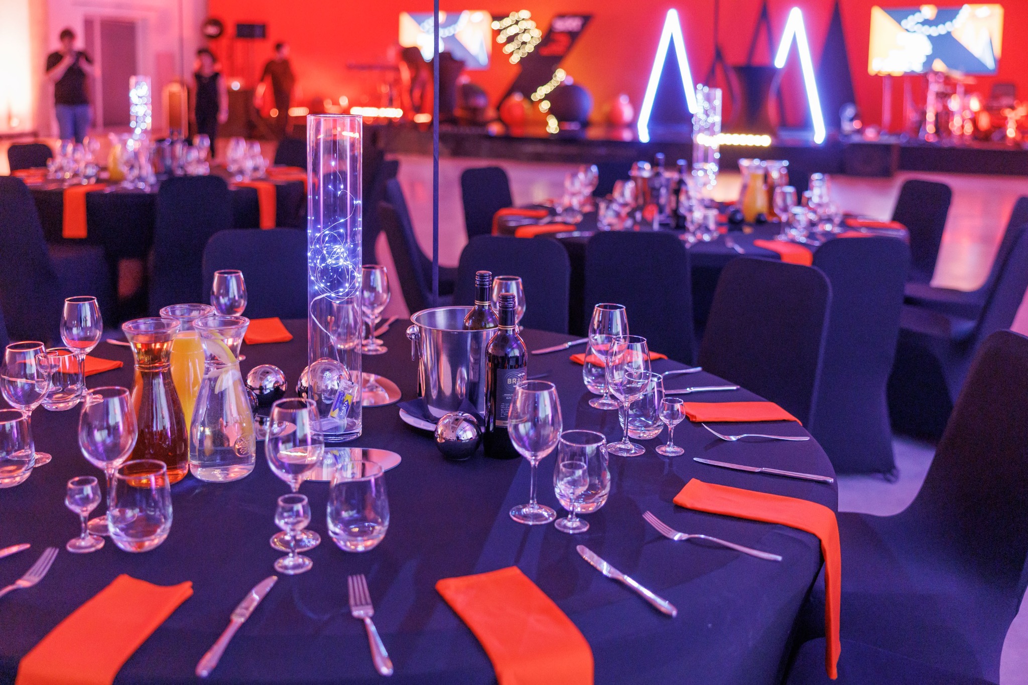 Uroczysty stół z czarnym obrusem, pomarańczowymi serwetkami, kieliszkami, karafkami z napojami, butelkami wina w wiaderku z lodem oraz dekoracyjnymi elementami, w tle scena z oświetleniem.
