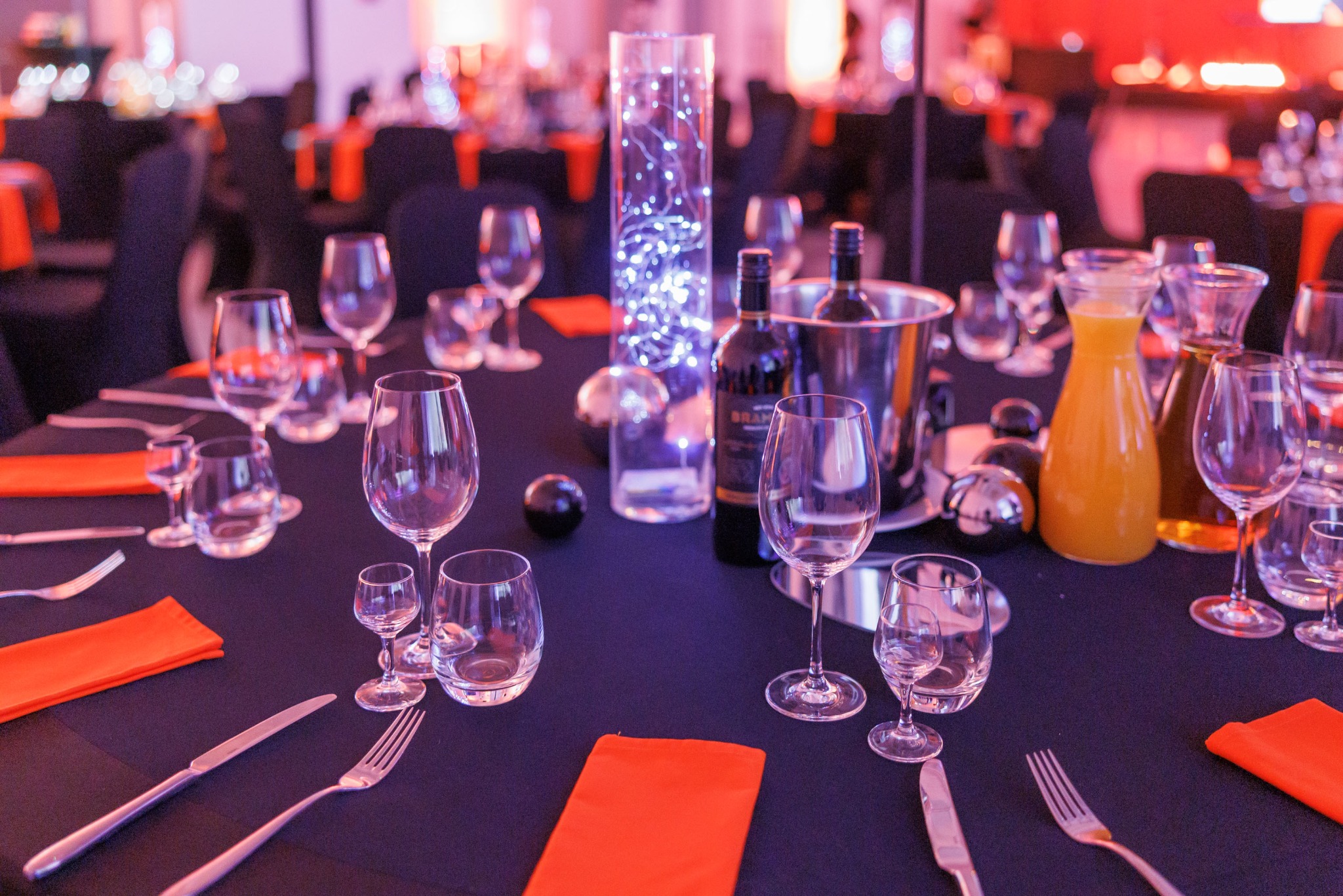 Uroczysty stół z czarnym obrusem, pomarańczowymi serwetkami, kieliszkami do wina, szklankami, sztućcami, butelkami wina w wiaderku z lodem i wysokim wazonem z lampkami LED, oświetlony fioletowym...