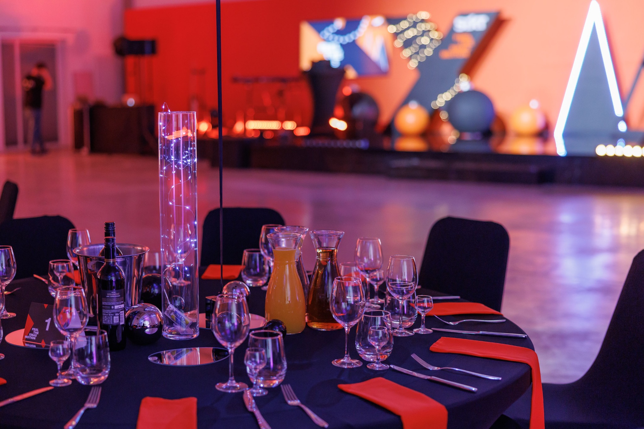 Uroczysty stół z czarnym obrusem i czerwonymi akcentami, nakryty szkłem, sztućcami i napojami, oświetlony czerwonym światłem, z dekoracjami scenograficznymi w tle.