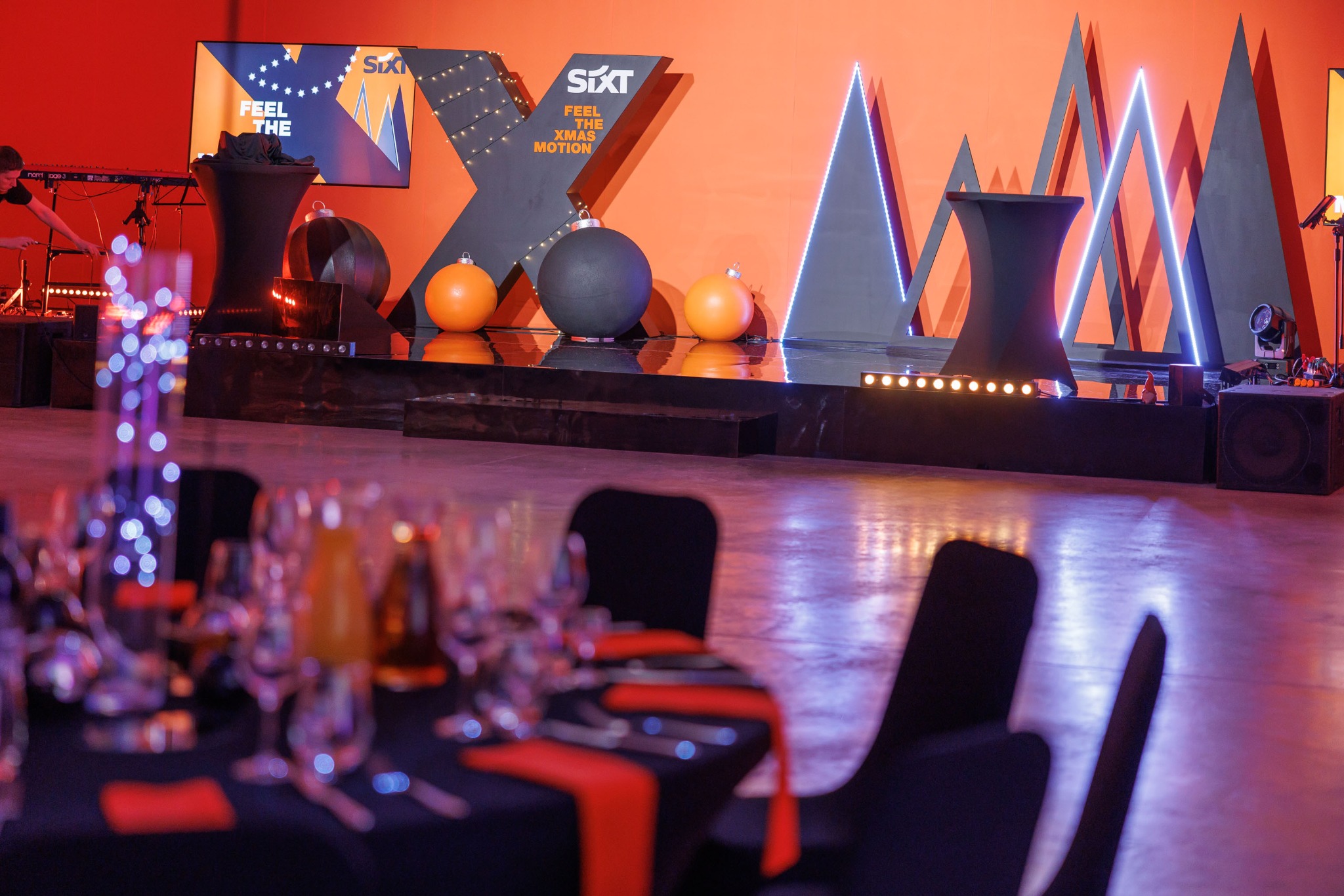 Scena z dekoracjami świątecznymi w postaci kul, podświetlanych trójkątów i logo firmy, widoczne nakryte stoły z kieliszkami i czerwonymi serwetkami, w tle muzyk grający na keyboardzie.