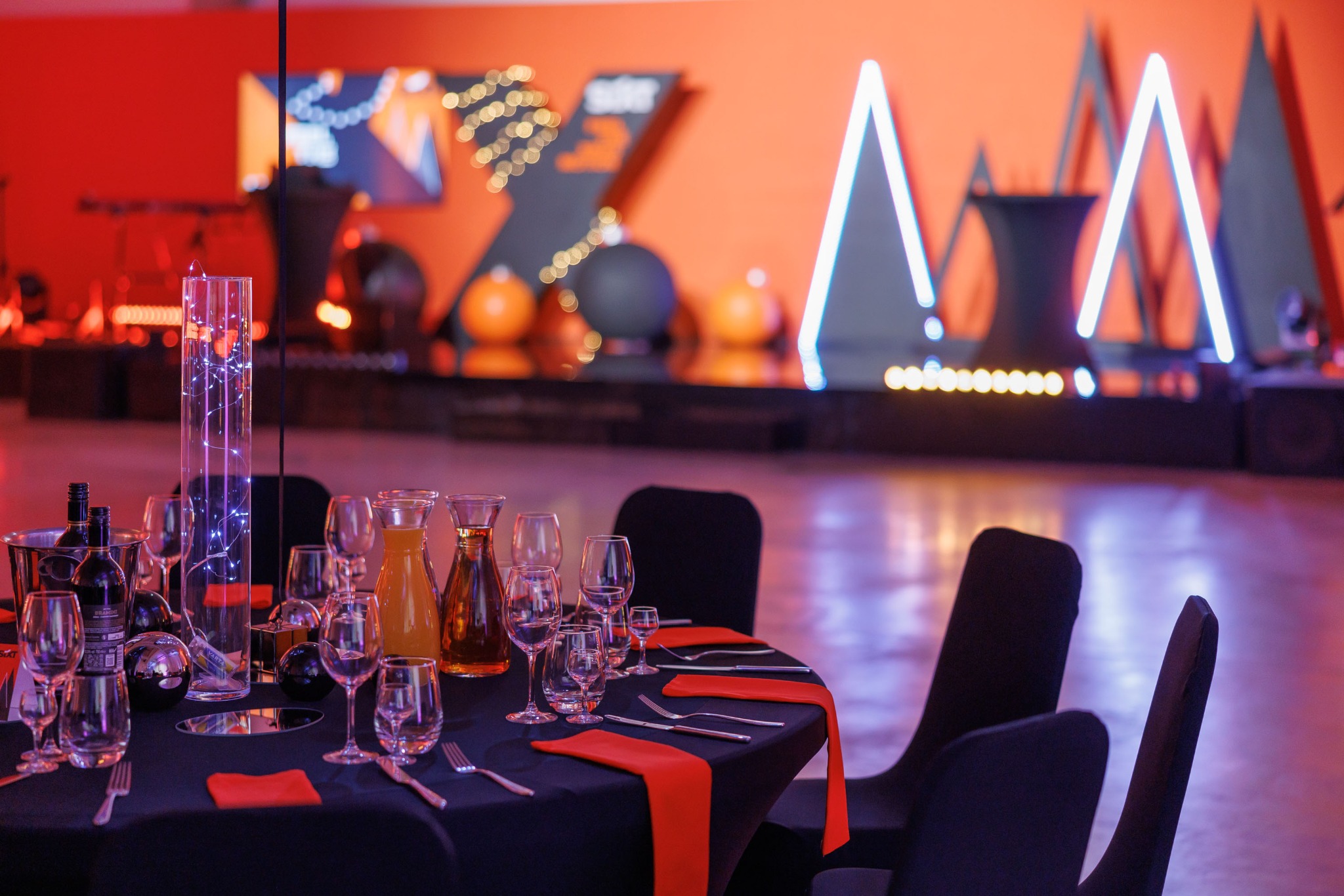 Elegancko nakryty stół na imprezie w Warszawie, z czarnym obrusem, czerwonymi serwetkami i szklanymi naczyniami, w tle dekoracje świetlne i scenografia w kolorach pomarańczowym i czarnym.