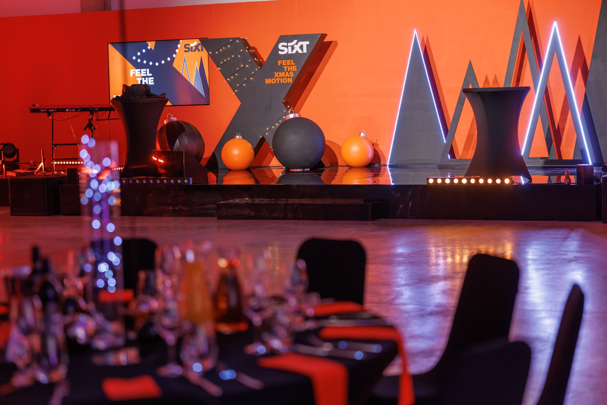 Scena z dekoracjami świątecznymi w pomarańczowo-czarnych barwach, logo firmy Sixt, stoły z nakryciami w tle, oświetlenie LED podkreślające geometryczne kształty.