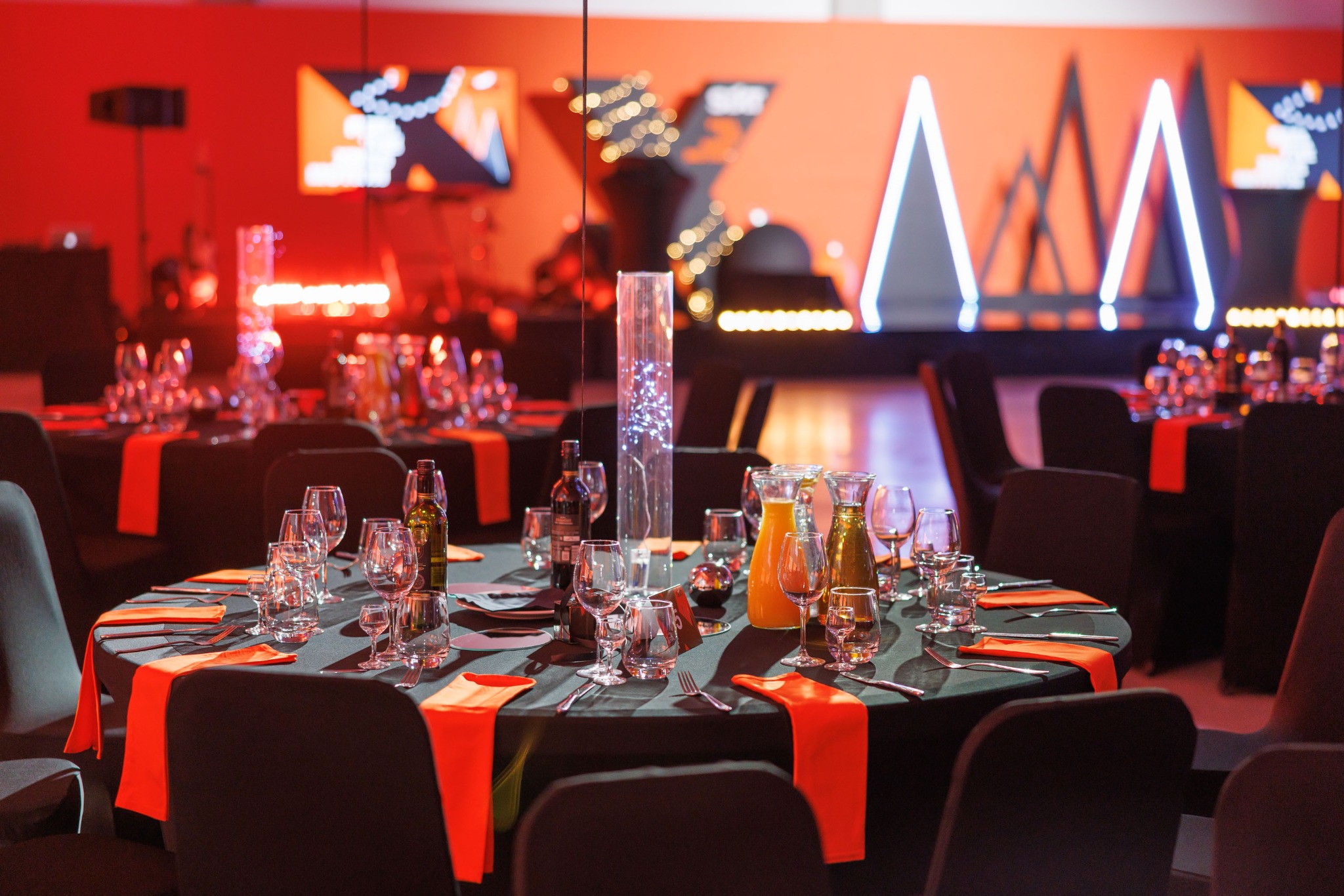Uroczysta aranżacja stołów z czarnymi obrusami i pomarańczowymi serwetkami, nakrytych szkłem i sztućcami, na tle sceny z geometrycznymi dekoracjami i oświetleniem w odcieniach czerwieni i pomarańczy.