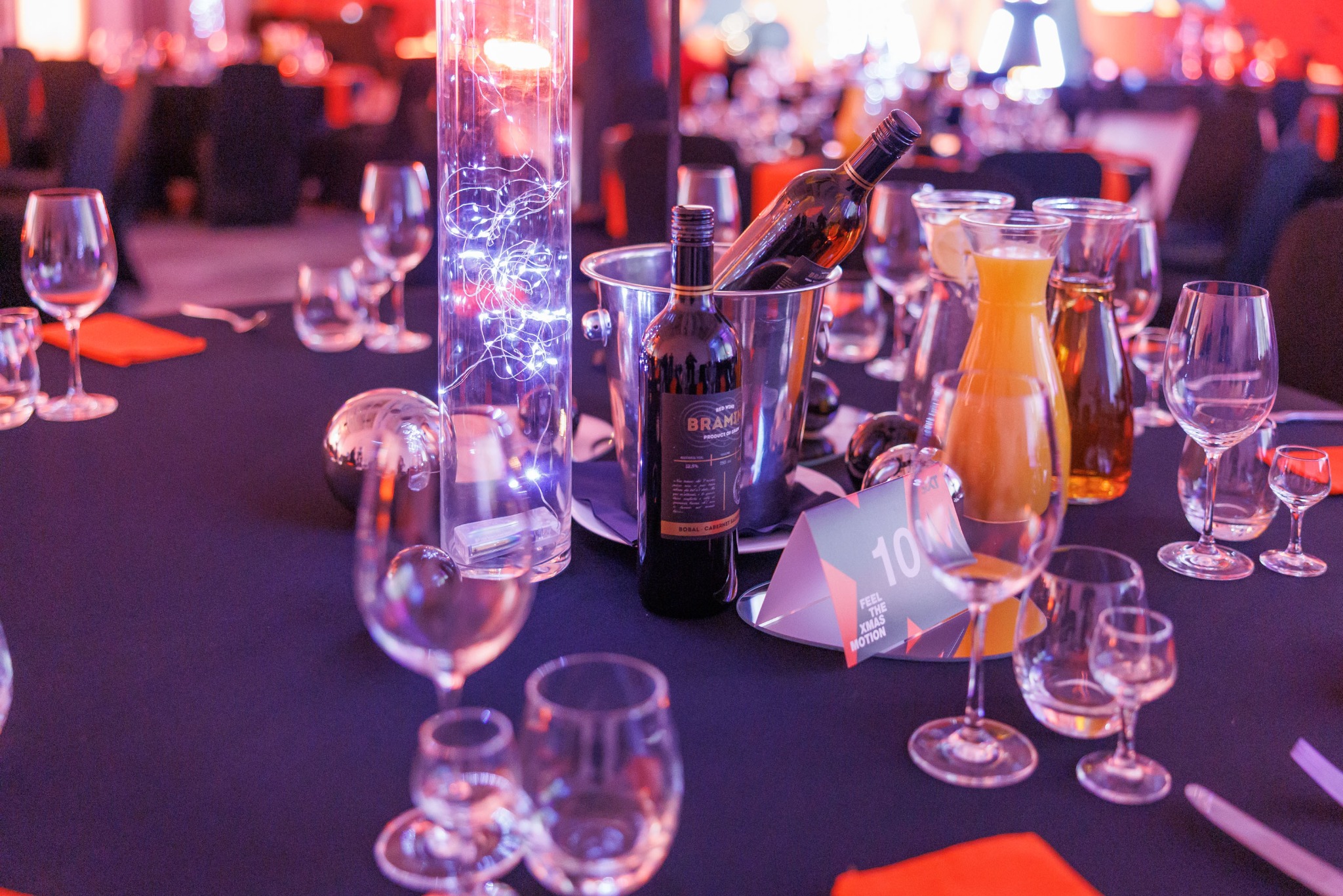 Uroczysty stół z czarnym obrusem, na którym stoją butelki wina w srebrnym wiaderku z lodem, karafki z sokami, szklanki, sztućce, srebrne kule dekoracyjne, wysoki szklany cylinder z lampkami LED...