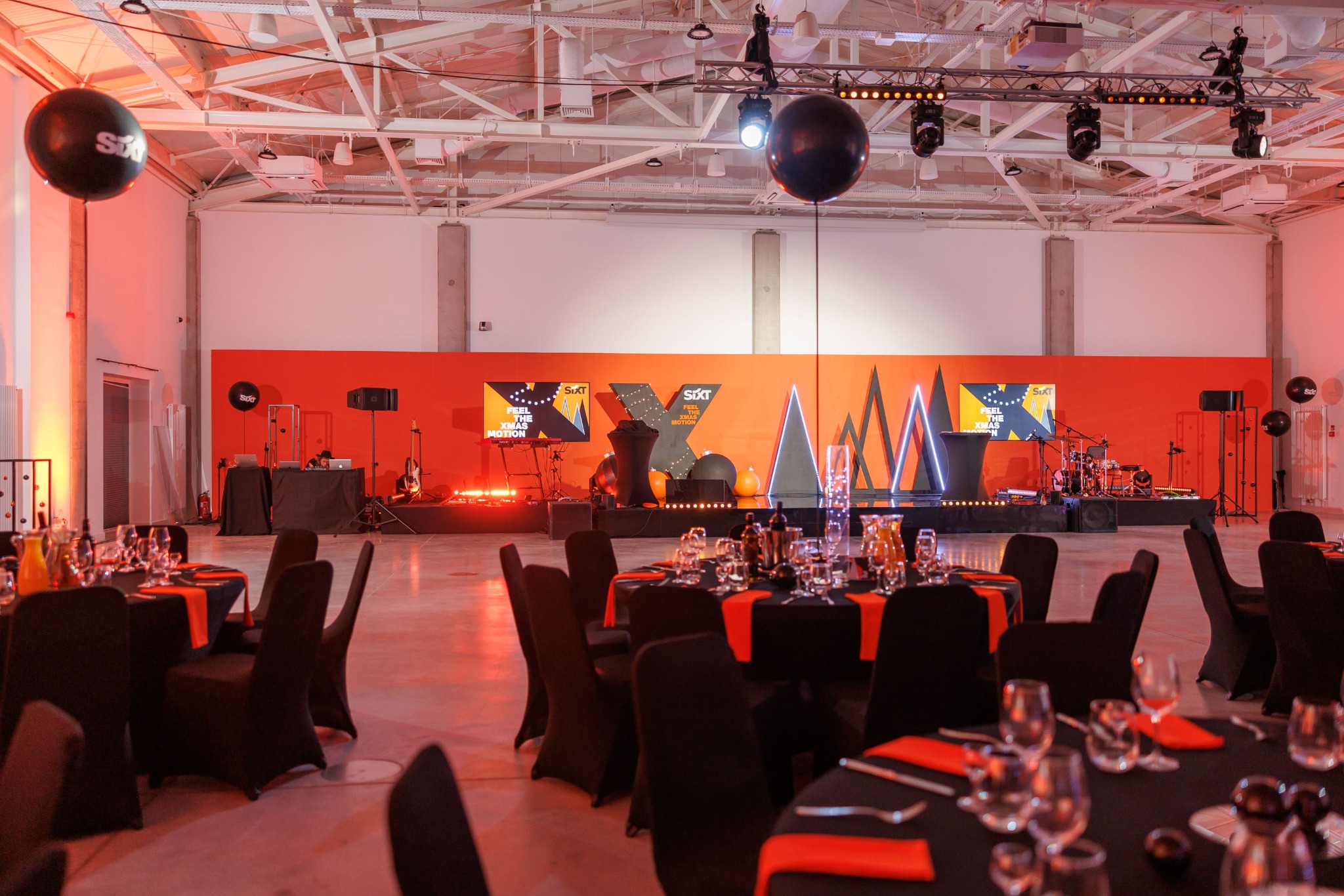 Sala konferencyjna przygotowana na imprezę firmową, stoły z czarnymi obrusami i pomarańczowymi serwetkami, scena z logo firmy, nagłośnienie i oświetlenie, balony z logo firmy.