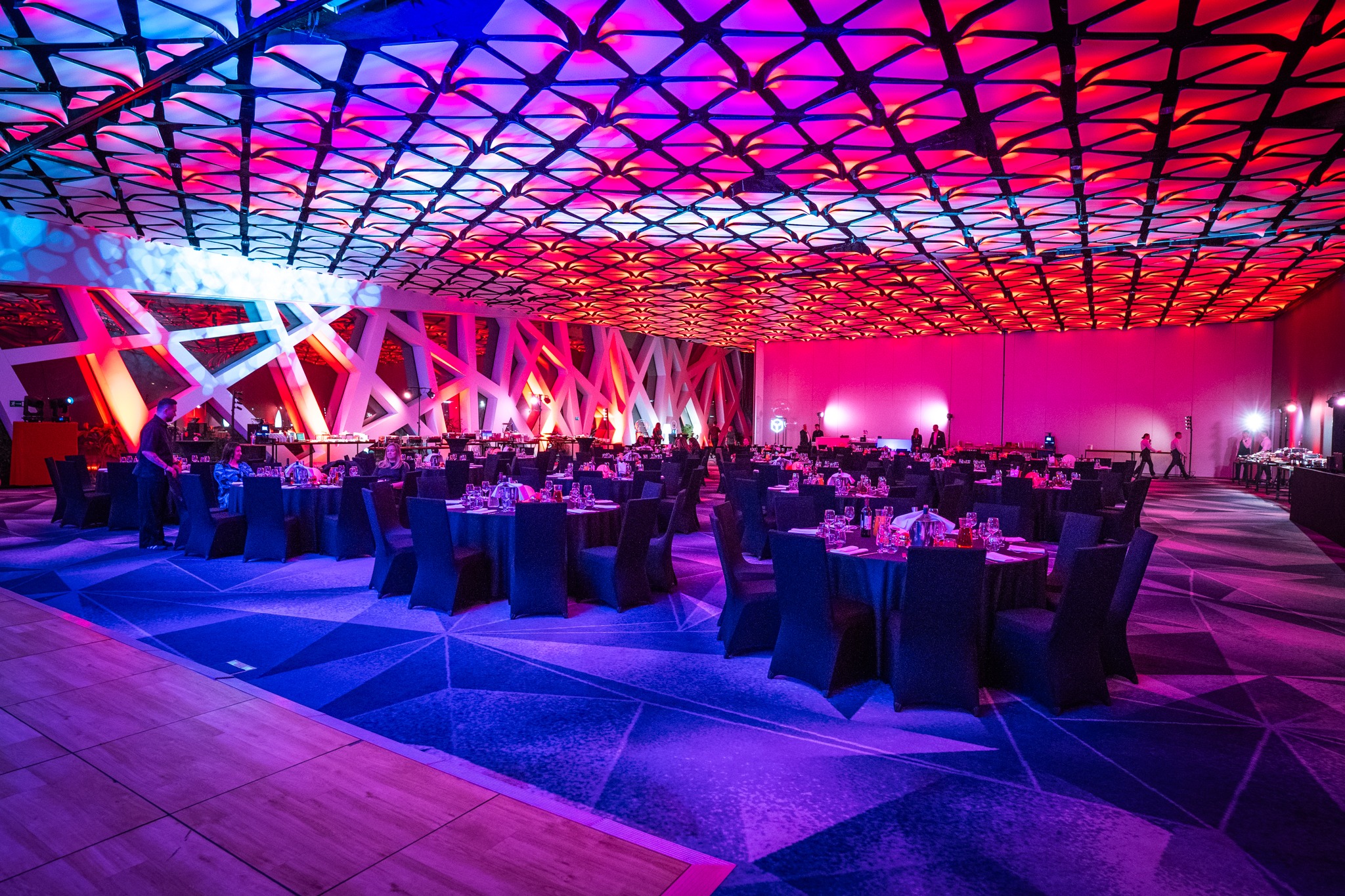 Sala bankietowa z okrągłymi stołami, nakryciami i krzesłami, oświetlona intensywnym, wielokolorowym światłem. Dekoracyjne oświetlenie sufitowe i geometryczne wzory na ścianach.