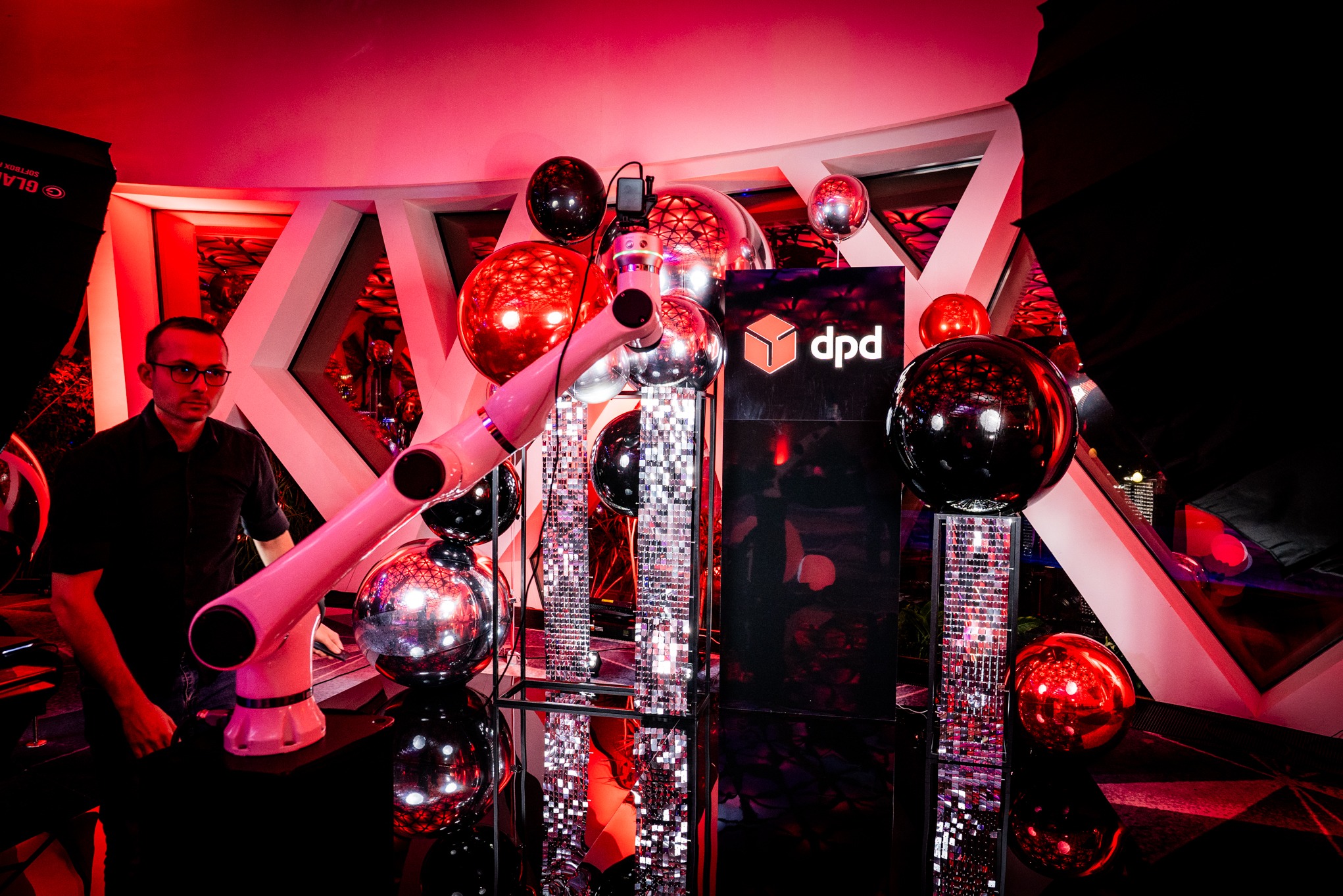 Event z robotem podającym prezenty DPD, dekoracje z balonów i błyszczących elementów. Mężczyzna w okularach obserwuje. Czerwone oświetlenie tworzy klubową atmosferę.