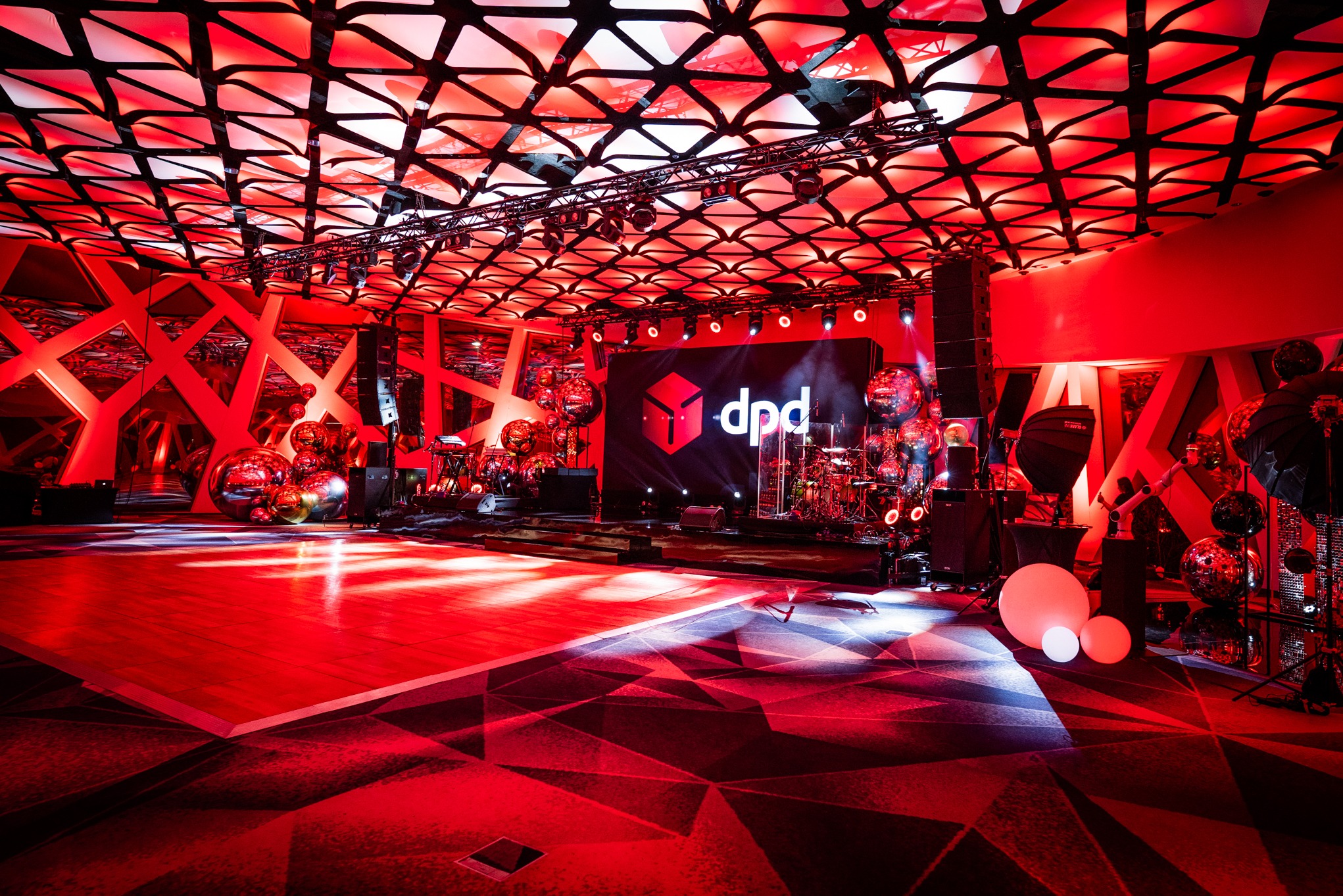 Czerwona sala eventowa z sceną, nagłośnieniem, oświetleniem i ekranem z logo dpd. Dekoracje z metalicznych kul. Parkiet taneczny i geometryczny wzór na podłodze.