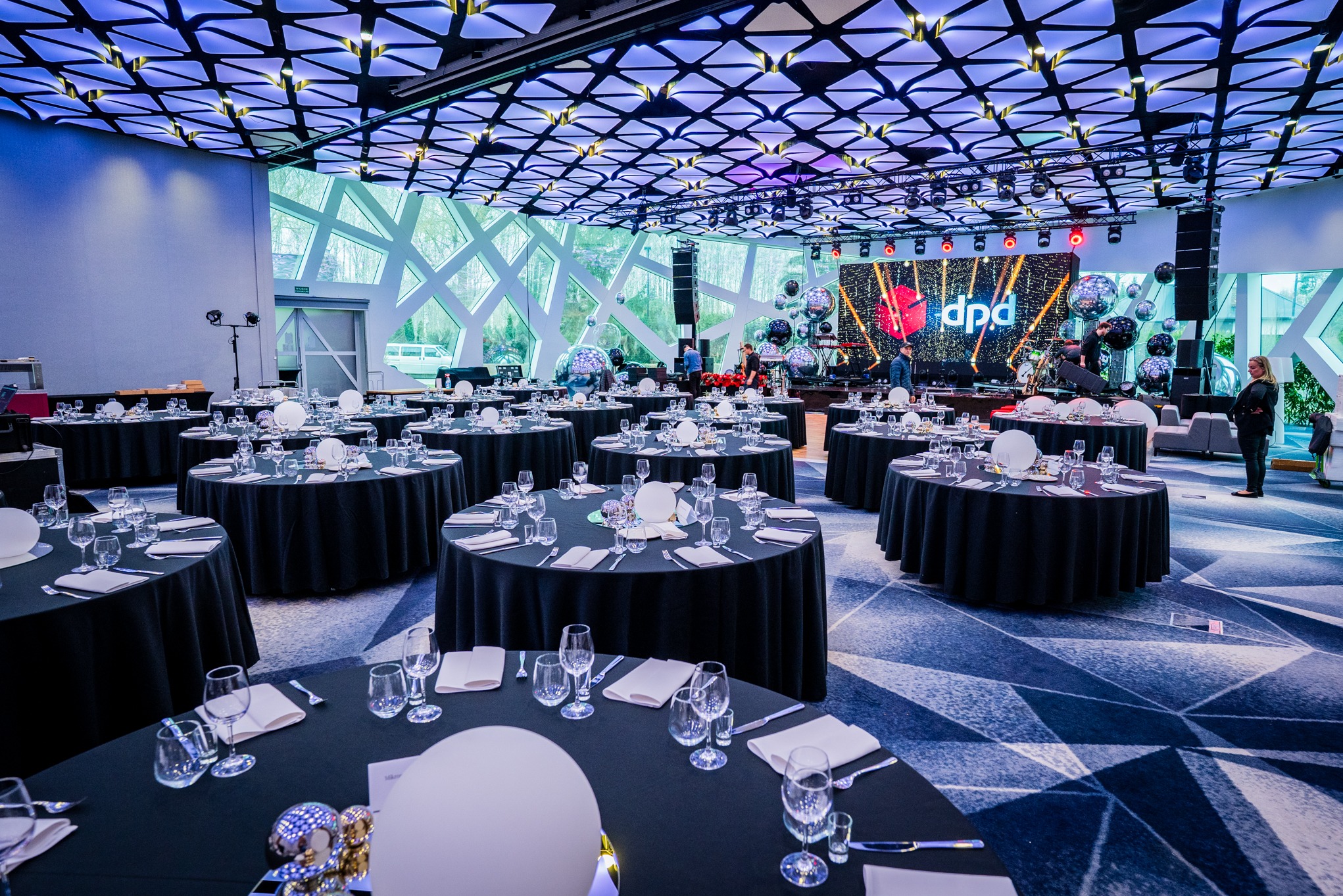 Sala bankietowa gotowa na imprezę firmową: okrągłe stoły z czarnymi obrusami, zastawa, scena z logo firmy DPD i dekoracje. Eleganckie oświetlenie sufitowe.