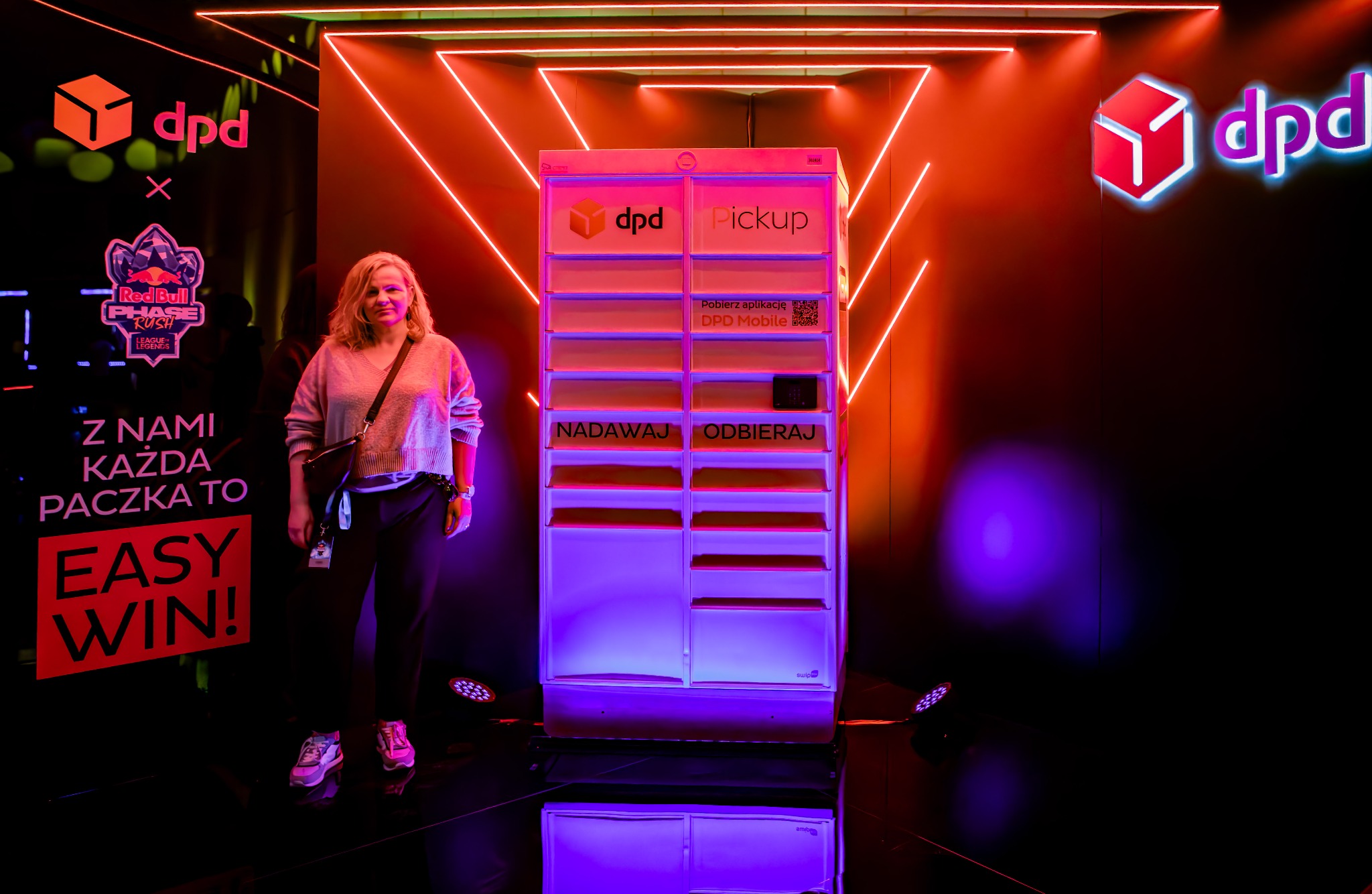Kobieta stoi obok paczkomatu DPD Pickup na tle neonowego logo DPD i Red Bull Phase Rush. Automat oświetlony na fioletowo, napisy 'Nadawaj' i 'Odbieraj'.